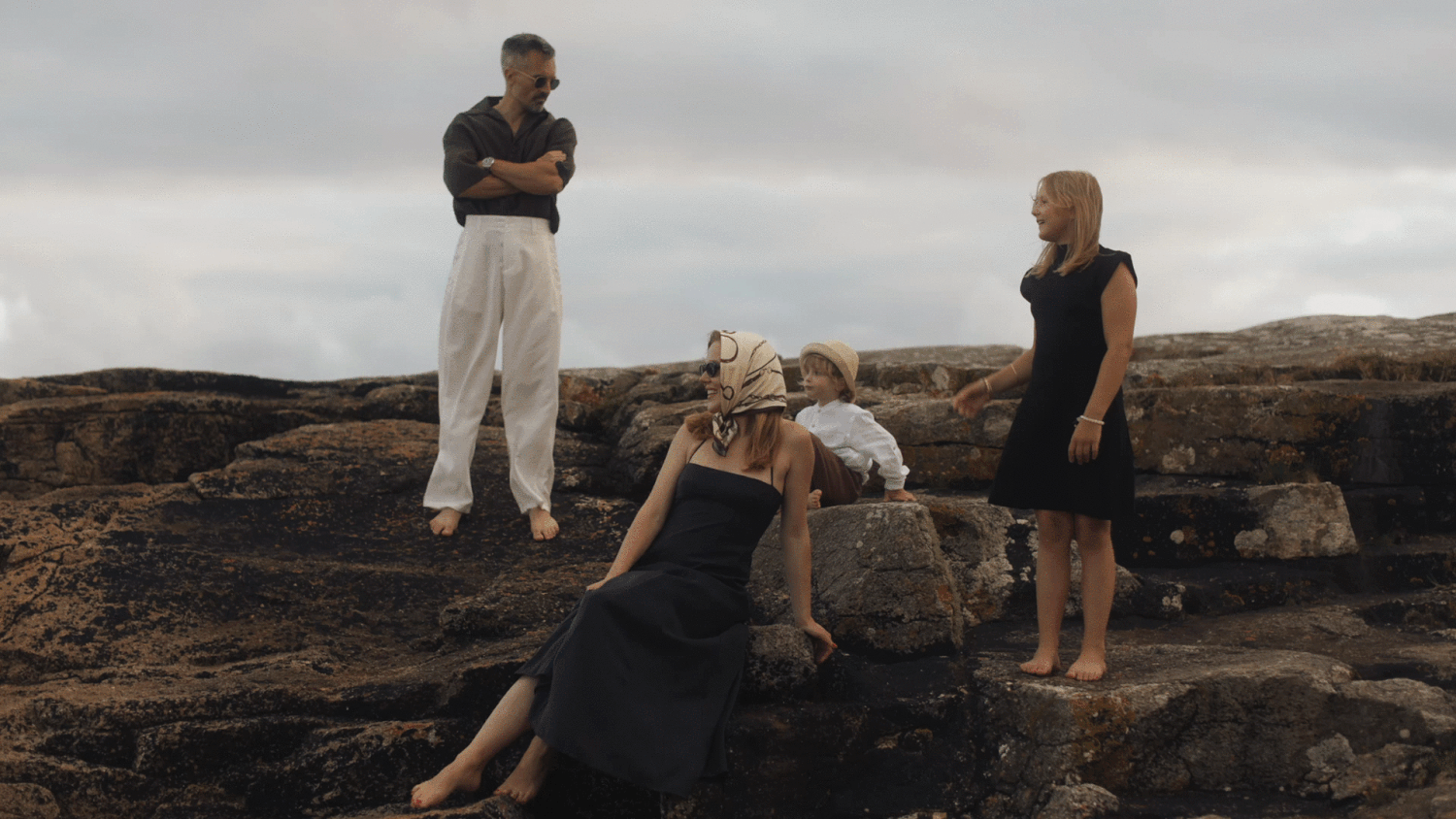 Family gathering on rocky coastal terrain, with adults and child posing outdoors under overcast skies.