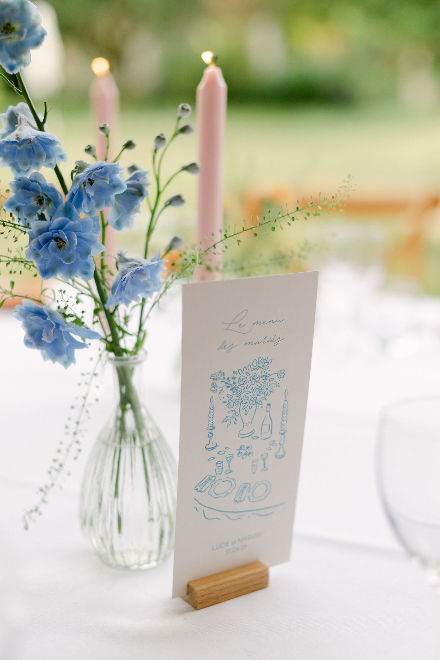 Élégante carte de table de mariage dans un support en bois à côté de fleurs de delphinium bleues et de bougies coniques roses sur une nappe