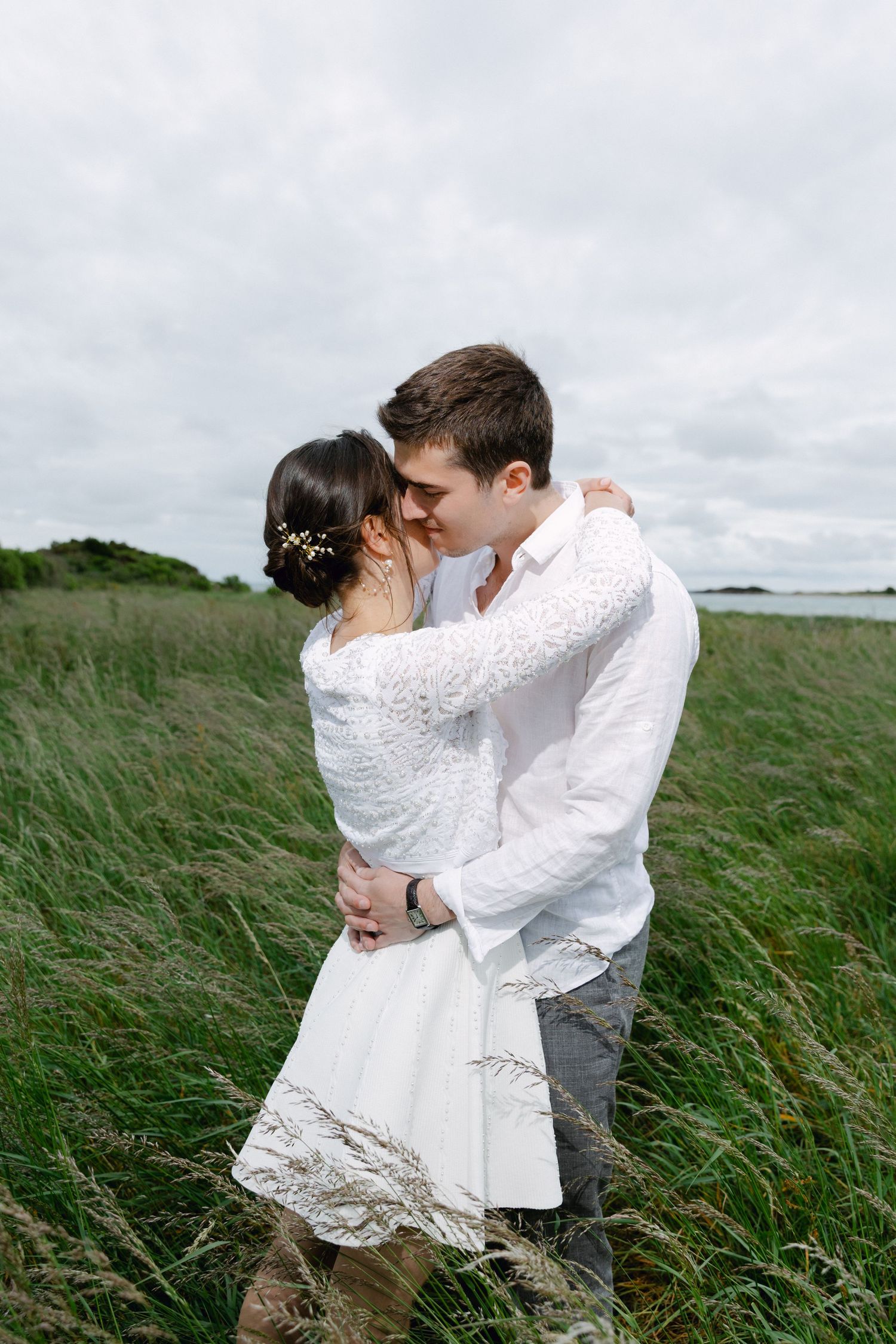 Un couple en tenue de mariage blanche s'embrasse et s'embrasse dans un champ vert luxuriant sous un ciel nuageux.