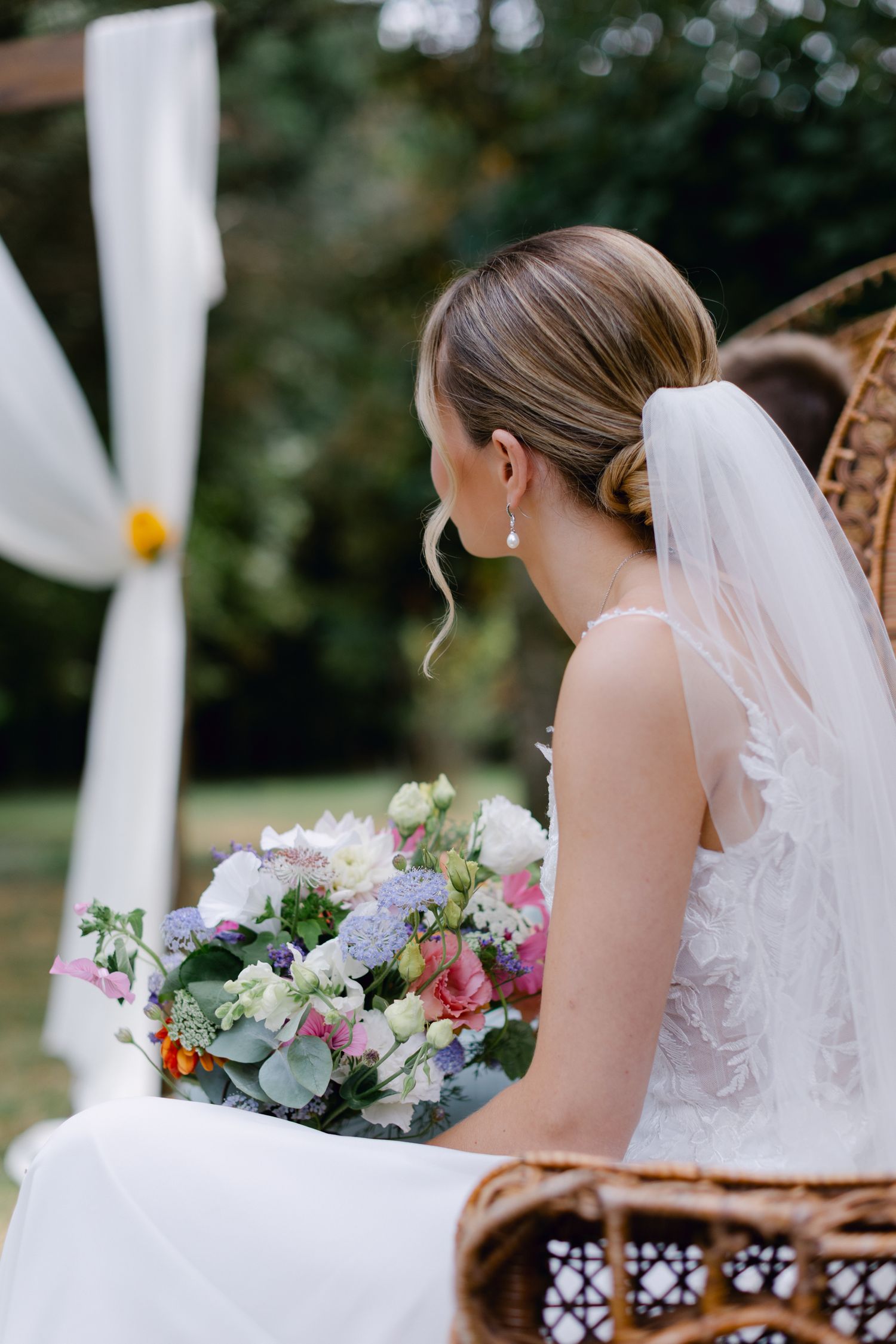 La mariée en robe de mariée en dentelle est assise avec un bouquet floral pastel près d'une arche drapée blanche.