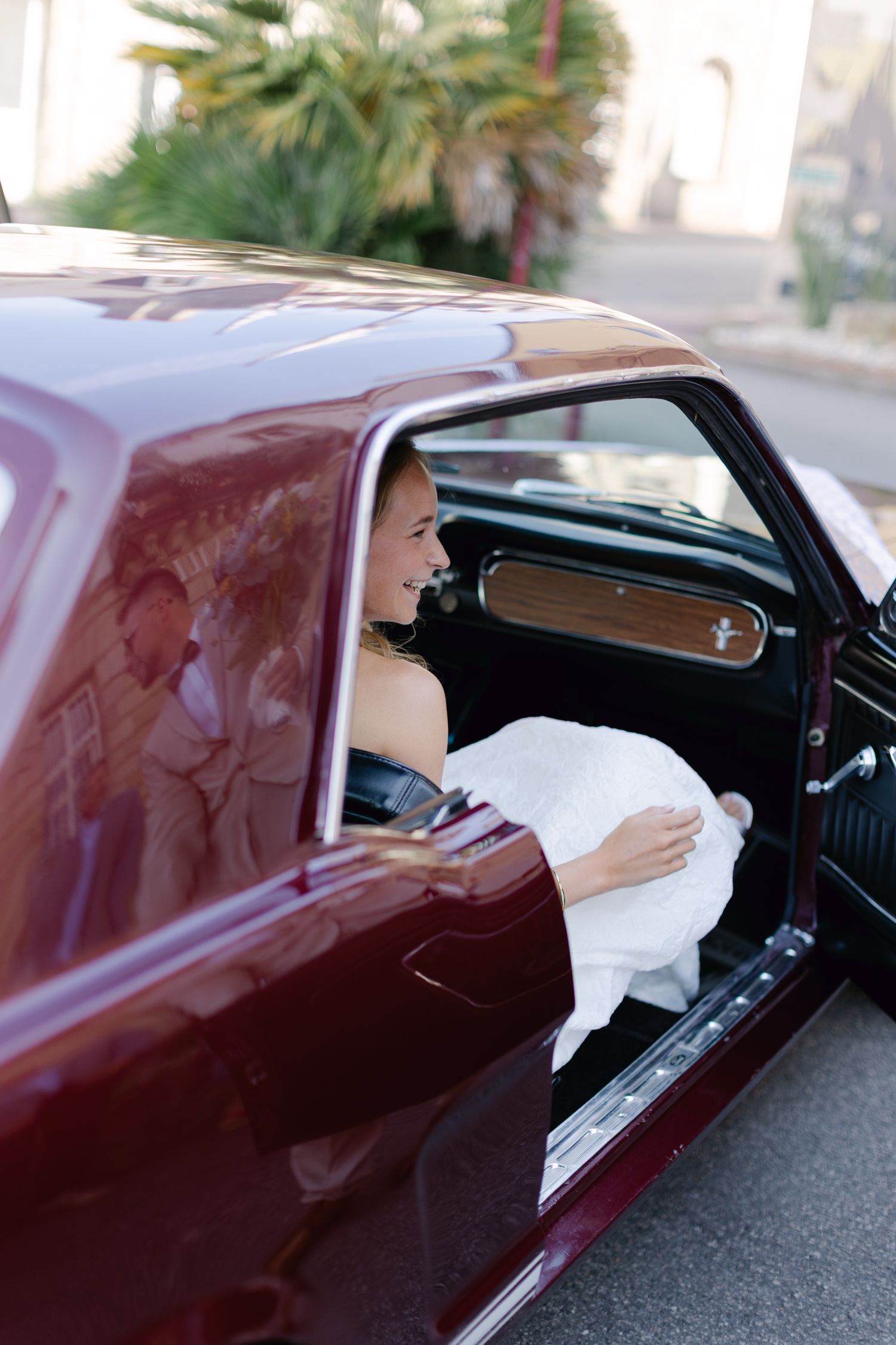 Mariée souriante en robe blanche assise dans une Ford Mustang vintage bordeaux le jour du mariage.