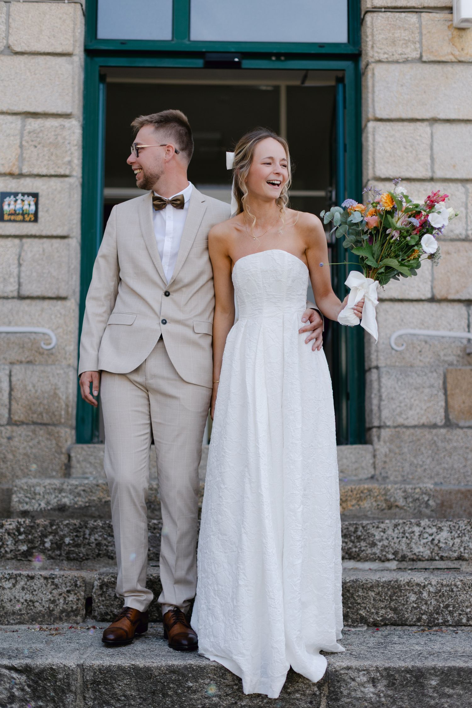 Mariée souriante en robe blanche sans bretelles tenant un bouquet coloré se tient avec le marié en costume beige sur des marches en pierre.