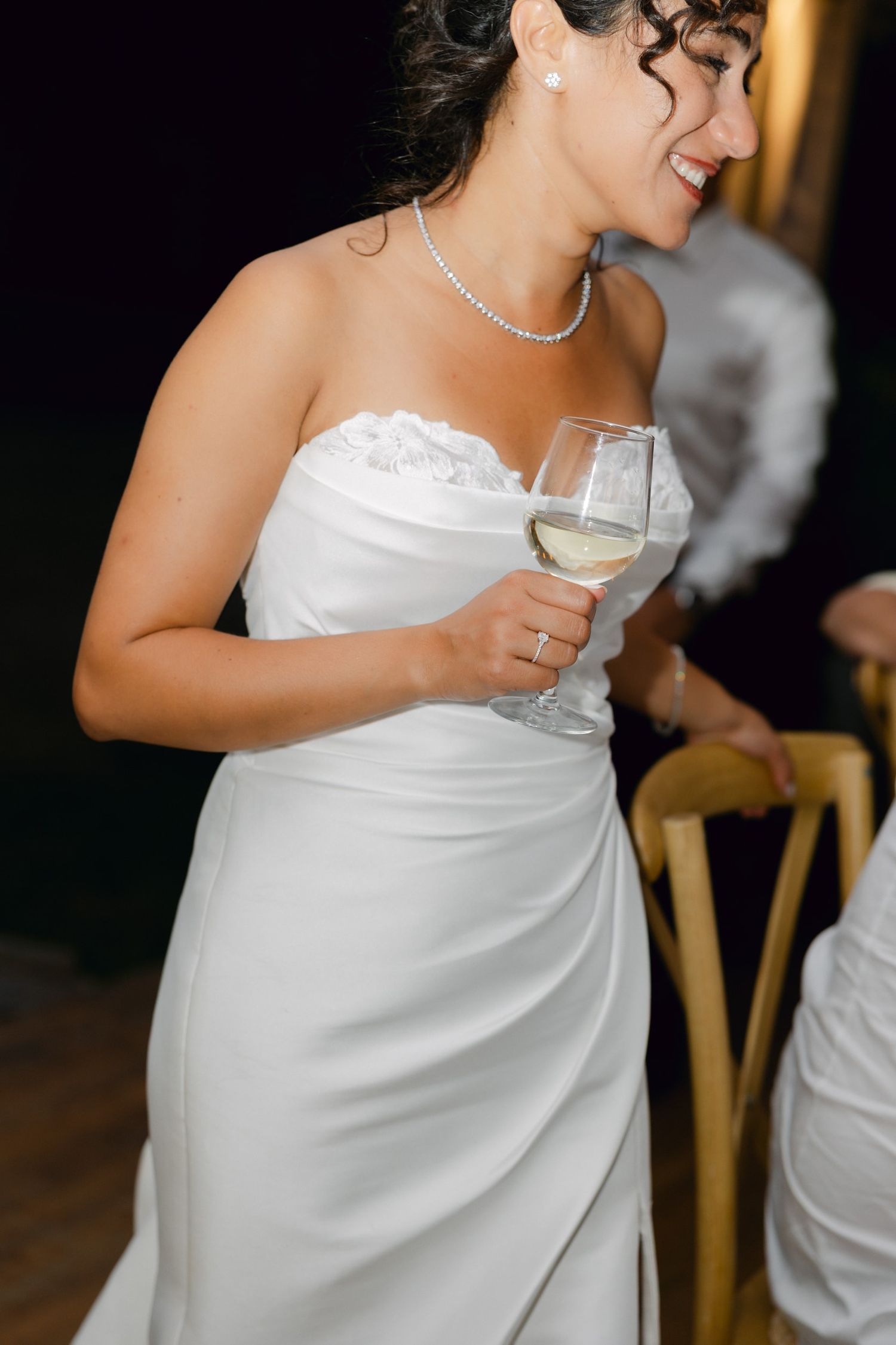 Smiling bride in strapless white gown holding a glass of white wine, wearing a diamond necklace and ring.