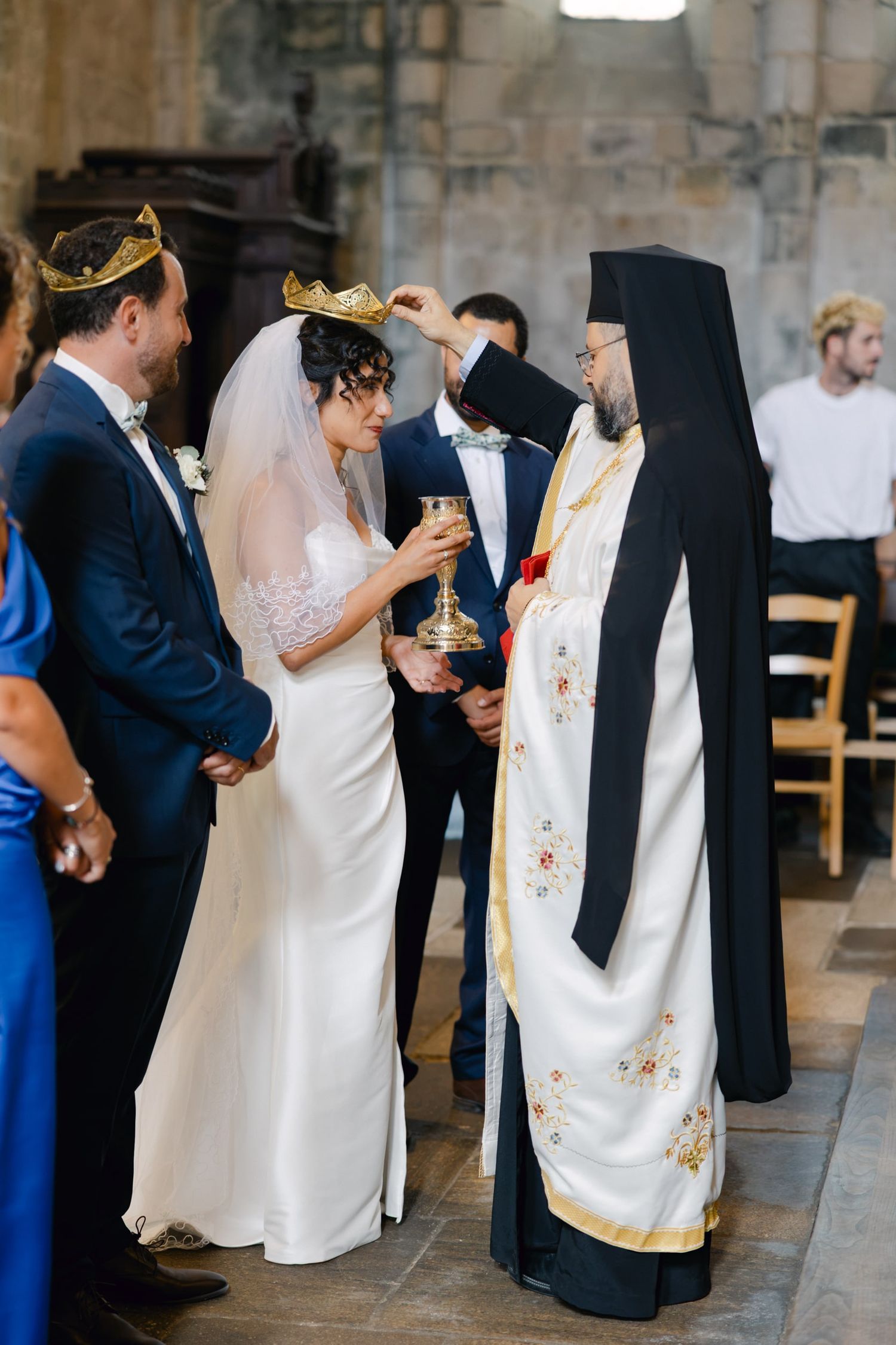 Orthodox priest placing crown on bride during traditional wedding ceremony with groom in blue suit.