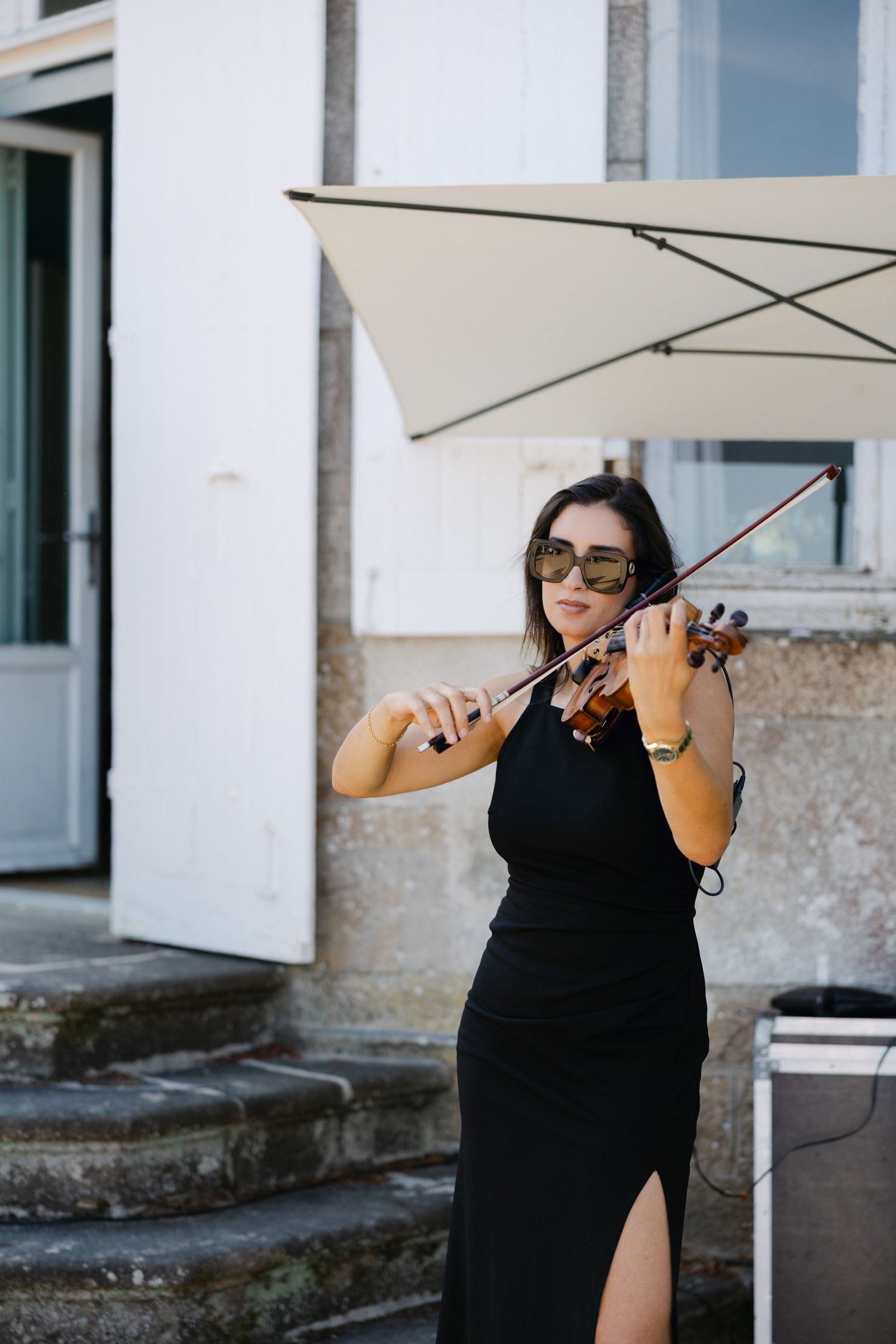 A woman in a black dress and sunglasses plays violin outdoors near building steps under a white umbrella.