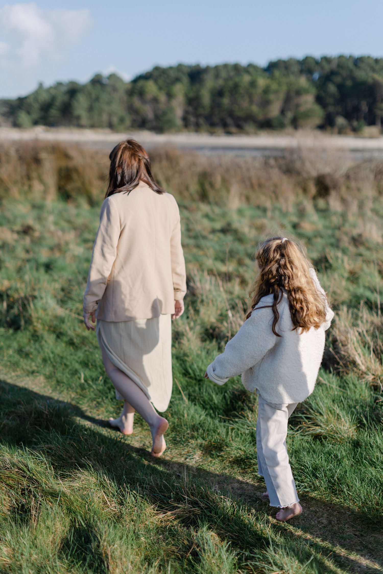 Une mère enceinte et sa jeune fille marchent pieds nus ensemble à travers une prairie côtière verte par derrière.
