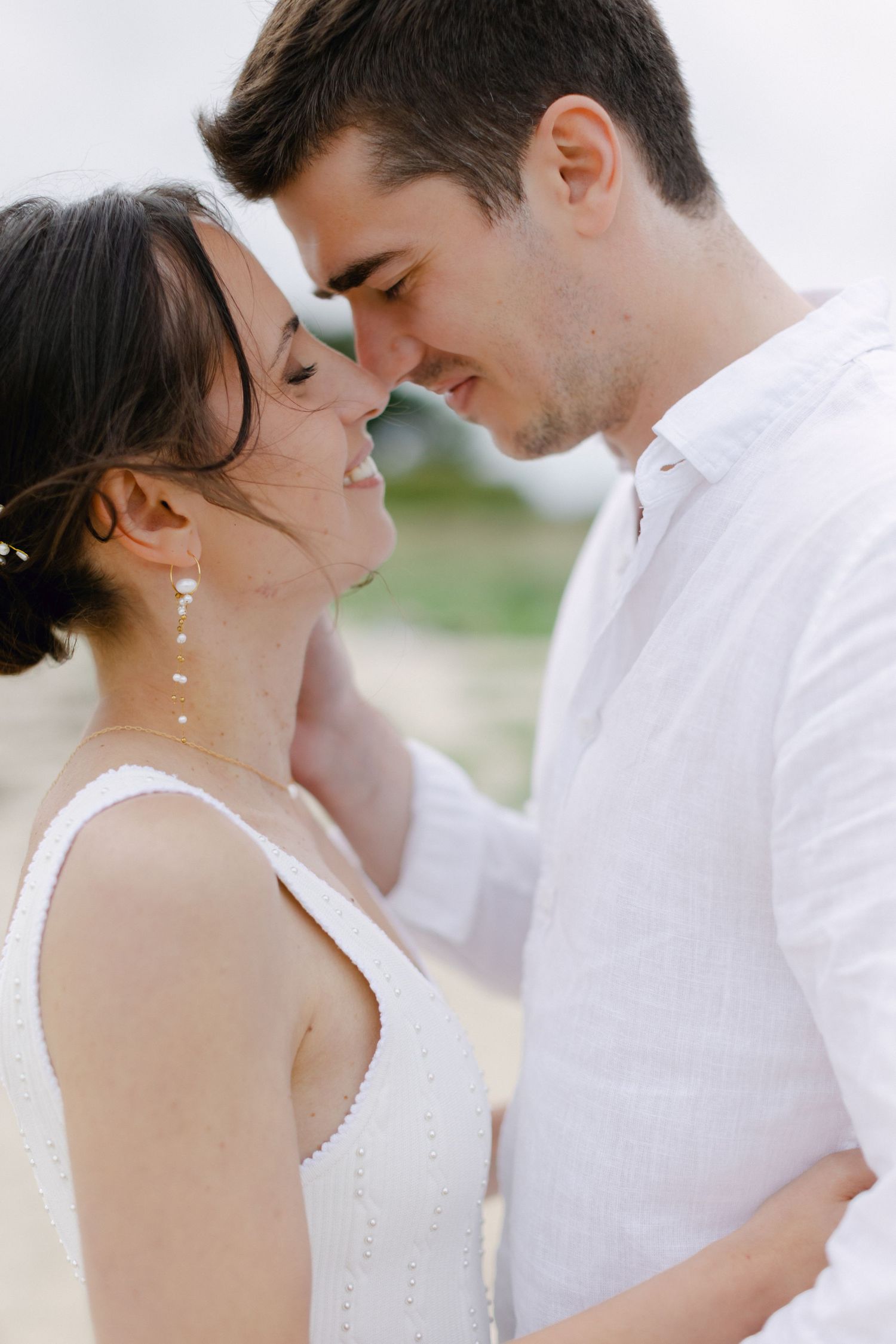 Un couple romantique en tenues blanches partageant un moment intime, les fronts presque touchants, souriant doucement à l'extérieur.