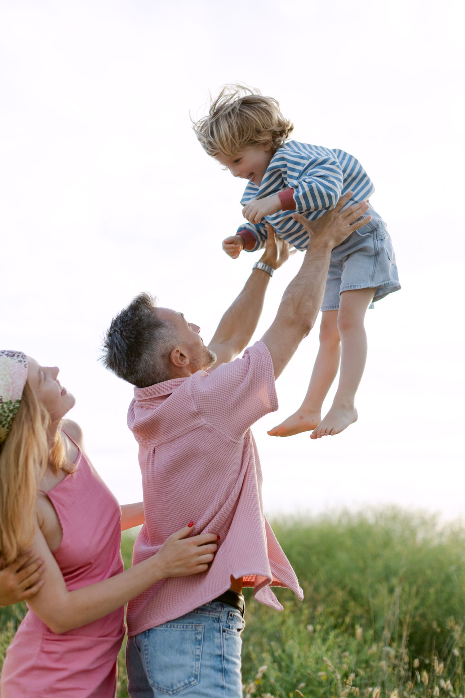 Le père soulève l'enfant souriant en l'air pendant que la mère regarde, profitant d'un moment familial ensoleillé en plein air.