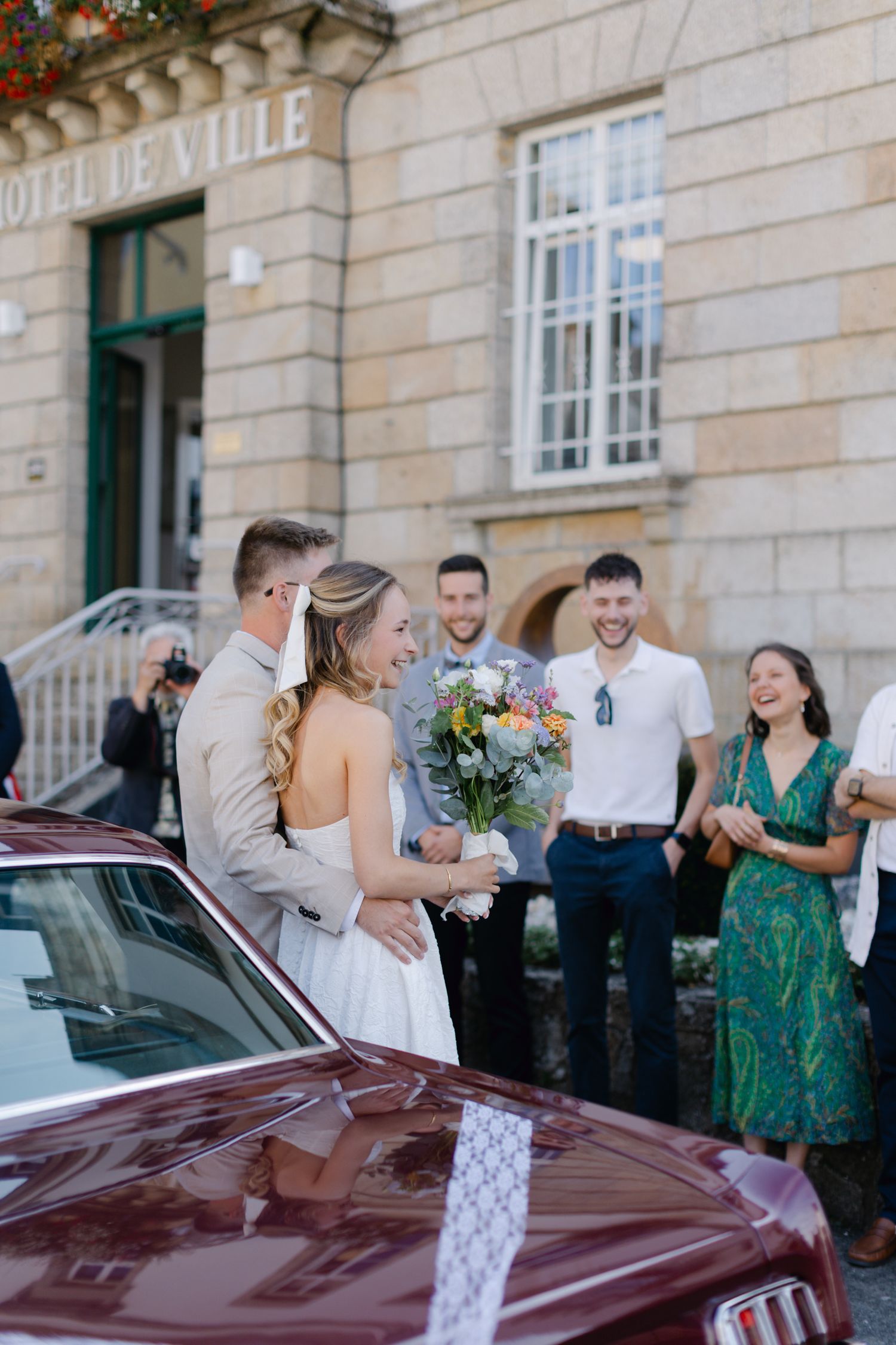 Une mariée souriante tenant un bouquet coloré embrasse le marié à l'extérieur de l'hôtel de Ville avec des invités de mariage à proximité.