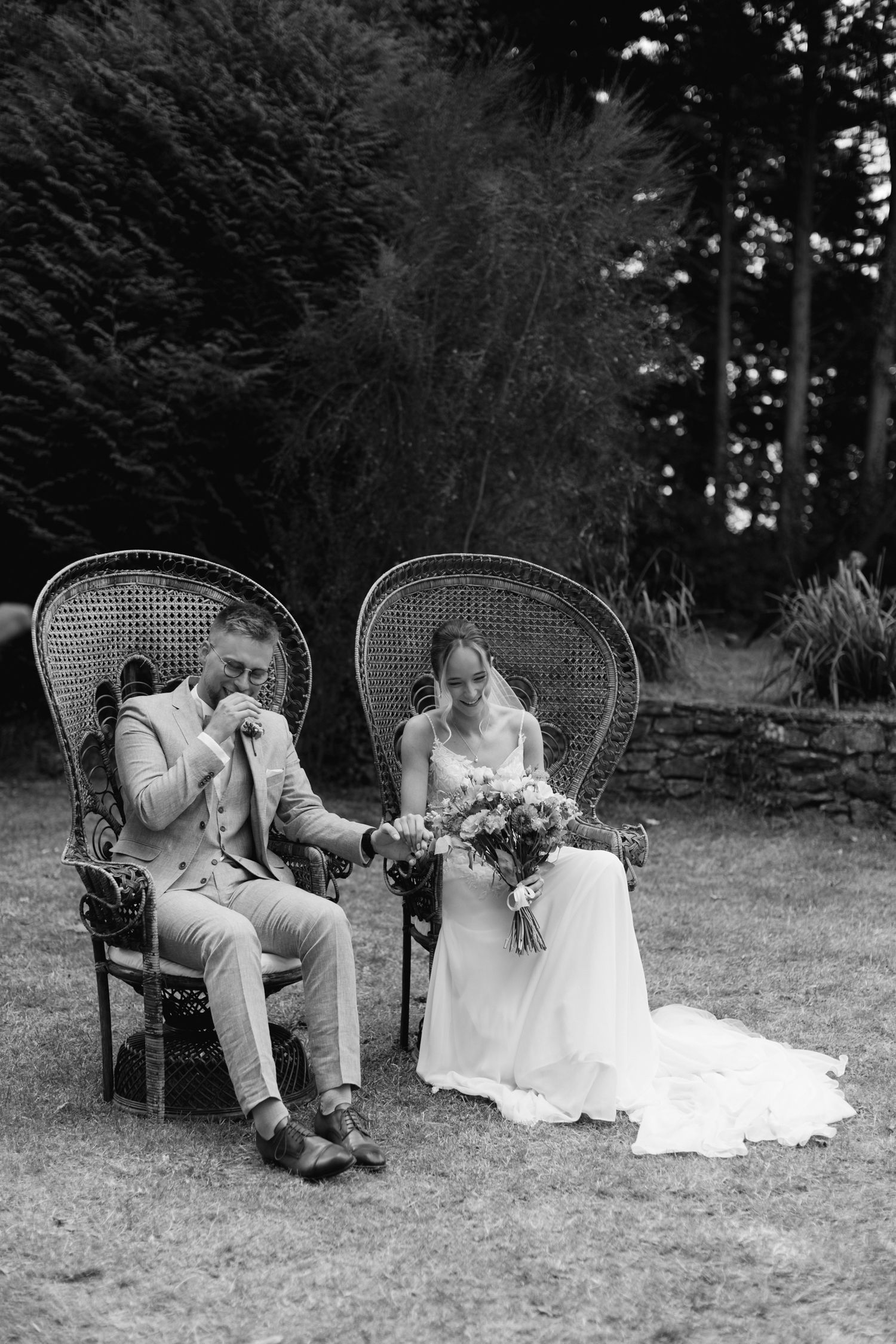 Photo en noir et blanc de la mariée et du marié assis dans des chaises en osier lors d'une cérémonie en plein air.