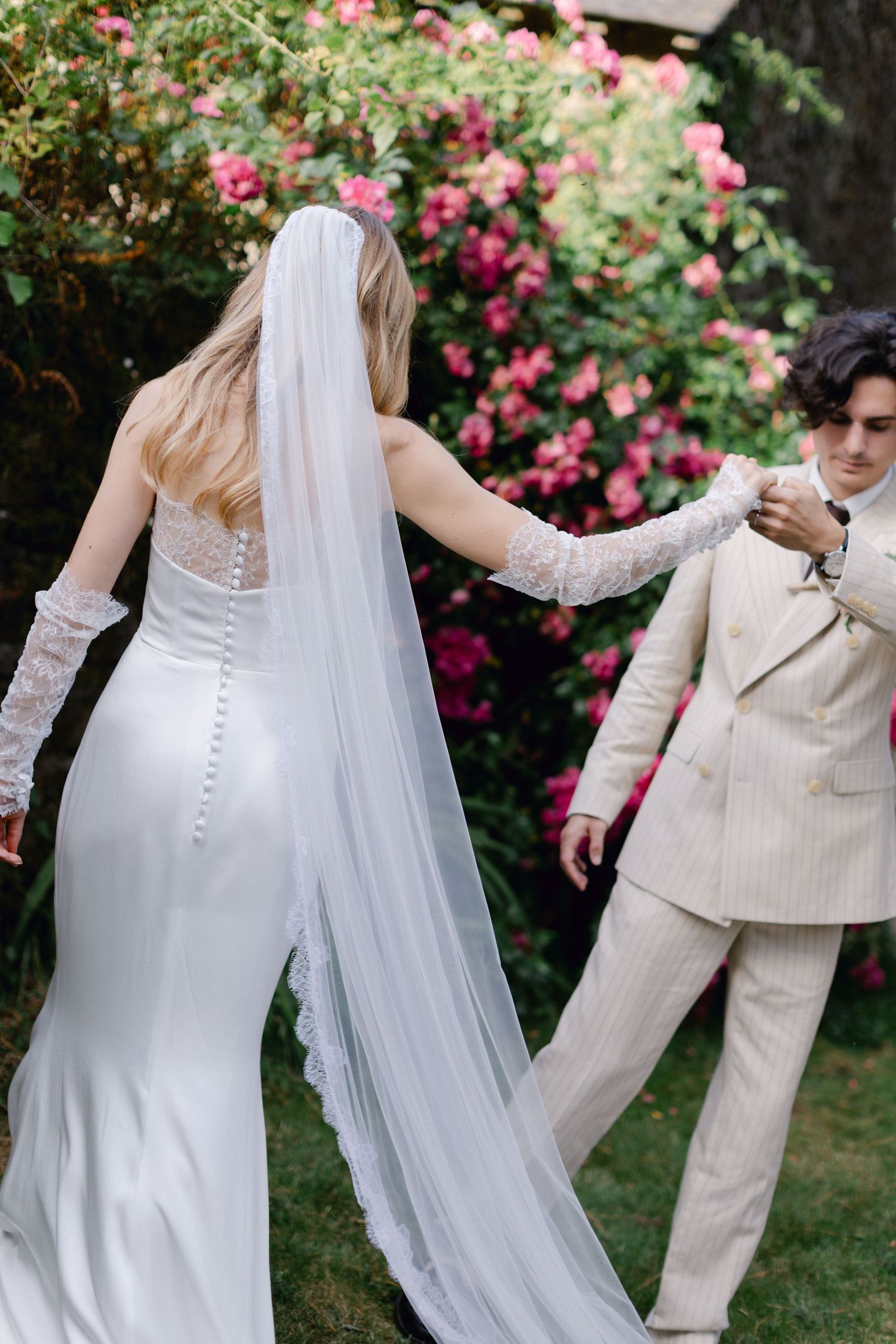 Bride in white gown with lace veil holding groom's hand near blooming pink roses outdoors.