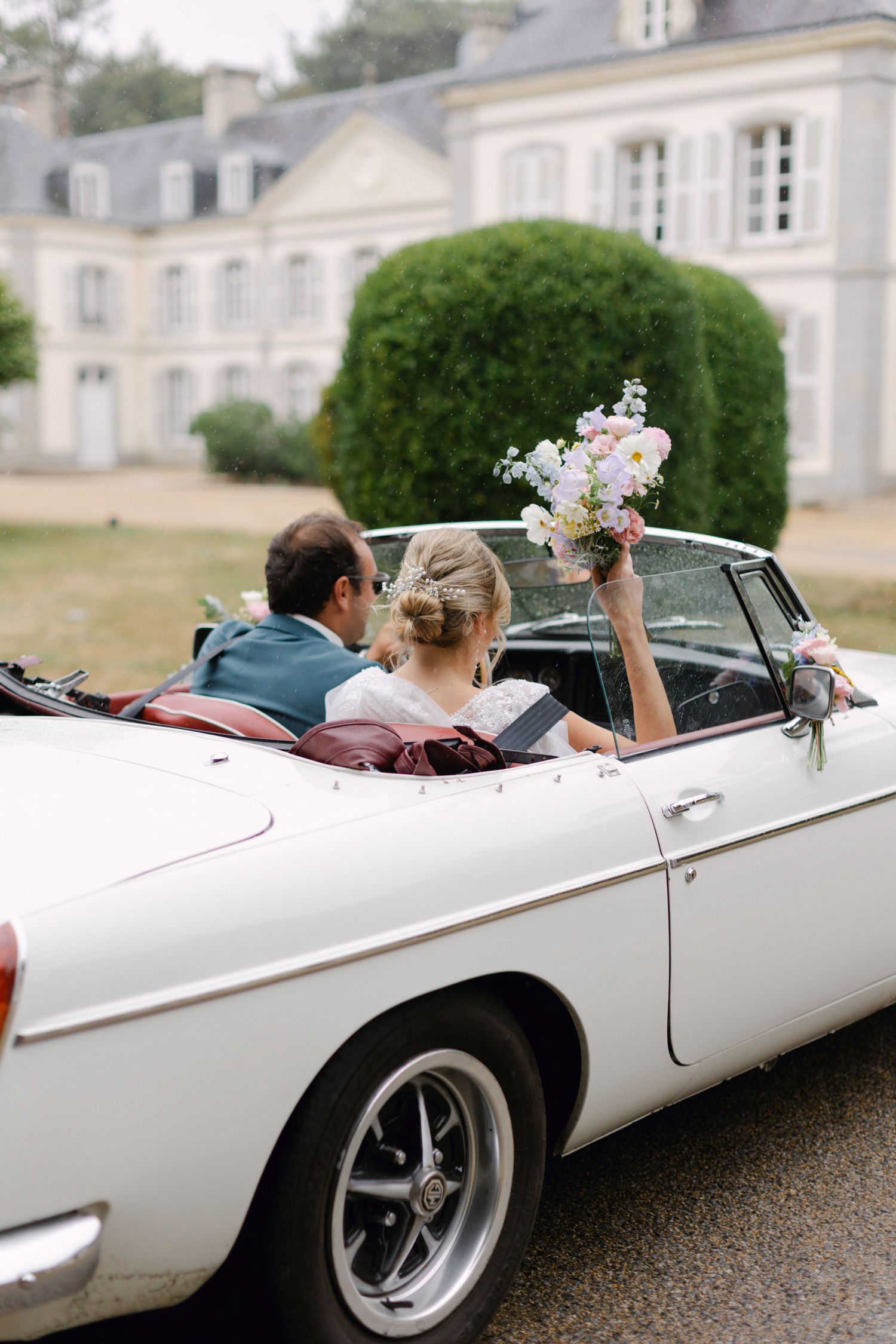 Les jeunes mariés montent dans une voiture décapotable vintage blanche devant un château français avec une mariée tenant un bouquet.