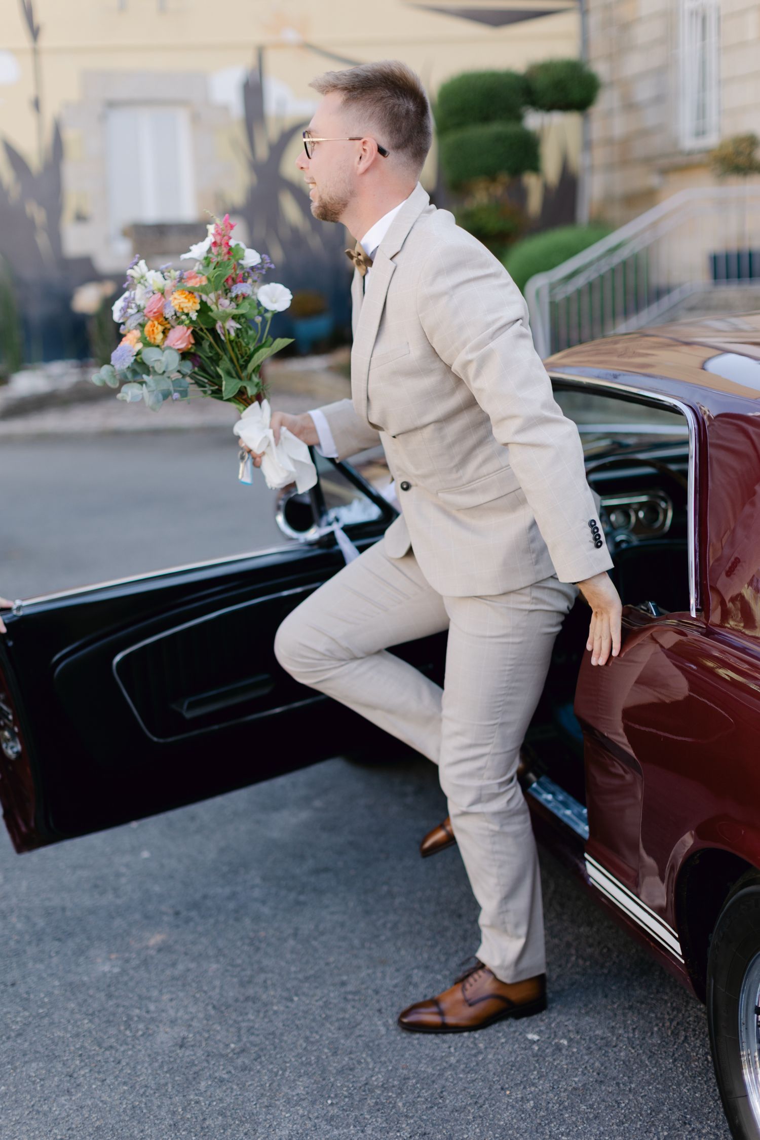 Marié en costume beige tenant un bouquet de mariée coloré sortant d'une voiture bordeaux vintage.