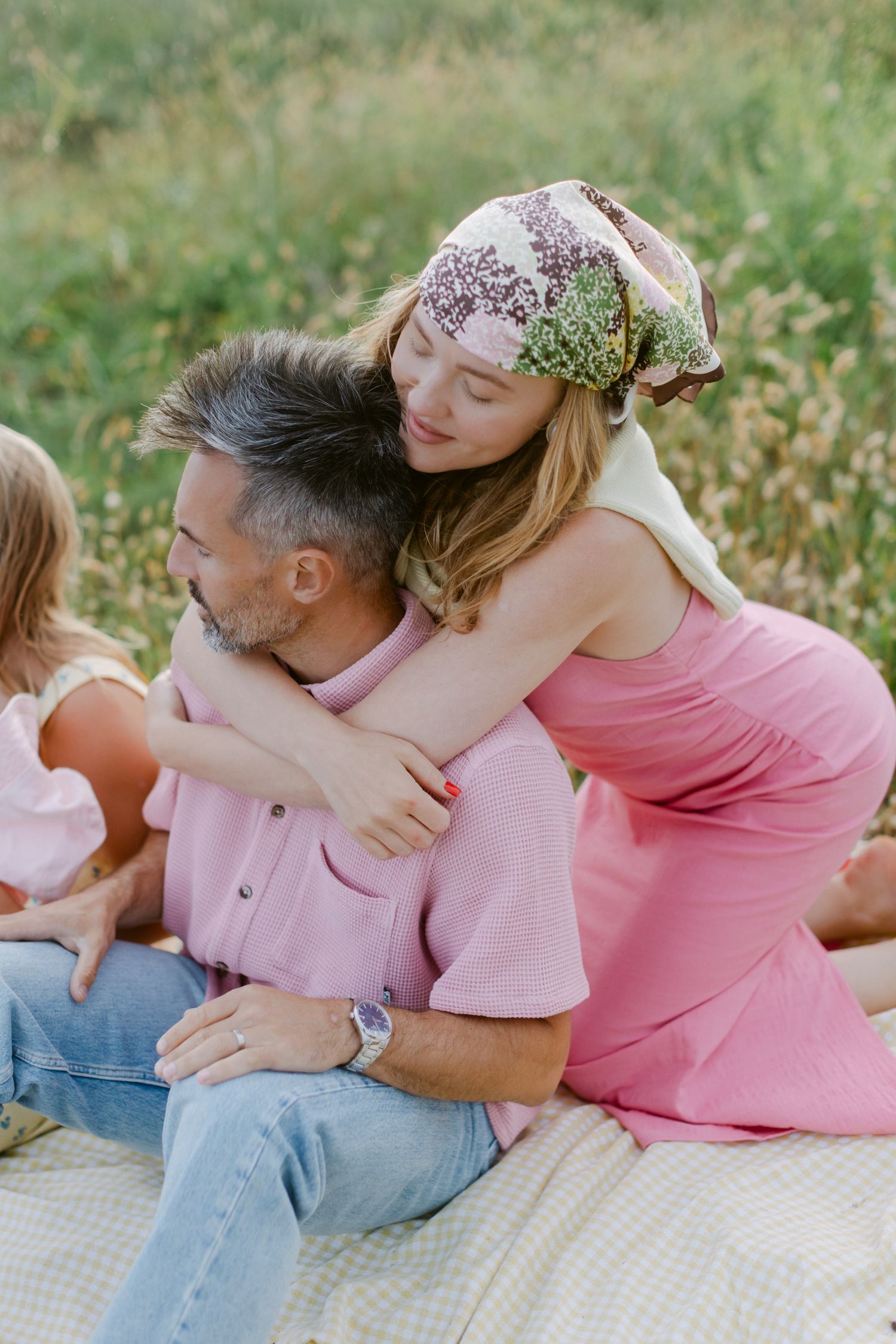 Une femme vêtue d'une robe rose et d'un foulard à fleurs embrasse un homme aux cheveux gris en chemise rose lors d'une photo de famille.