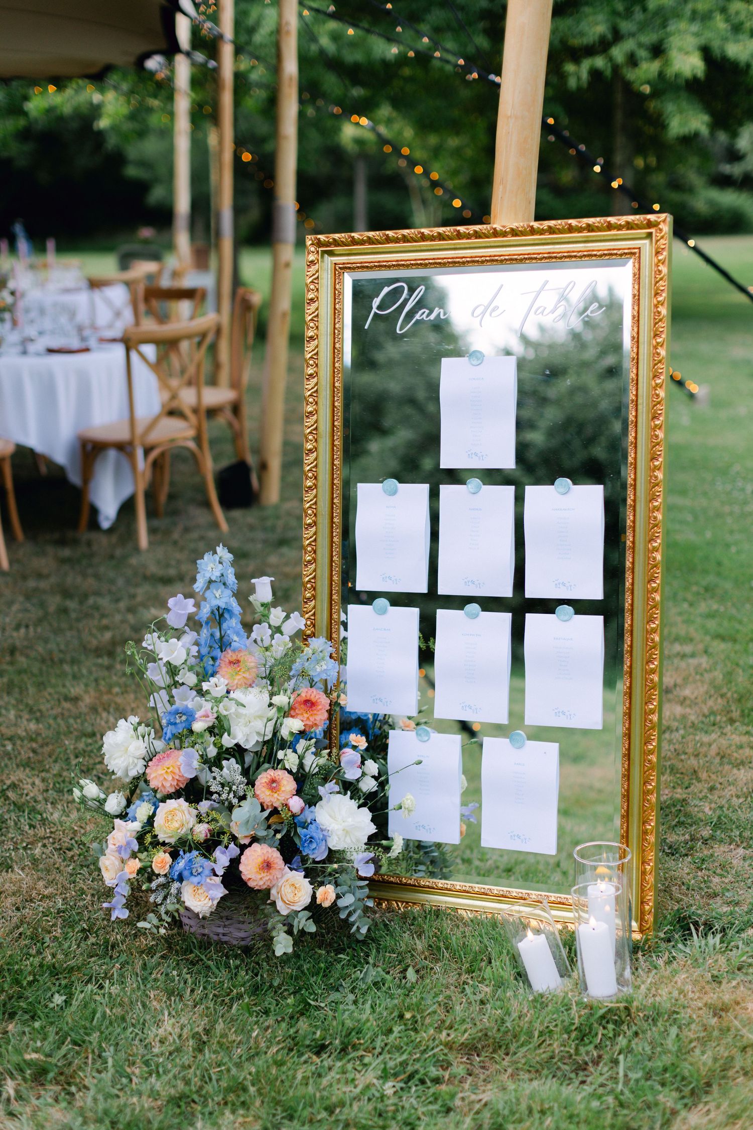 Tableau des sièges miroir encadré d'or étiqueté Plan de Table affiché sur l'herbe avec un arrangement floral bleu et pêche lors d'un mariage