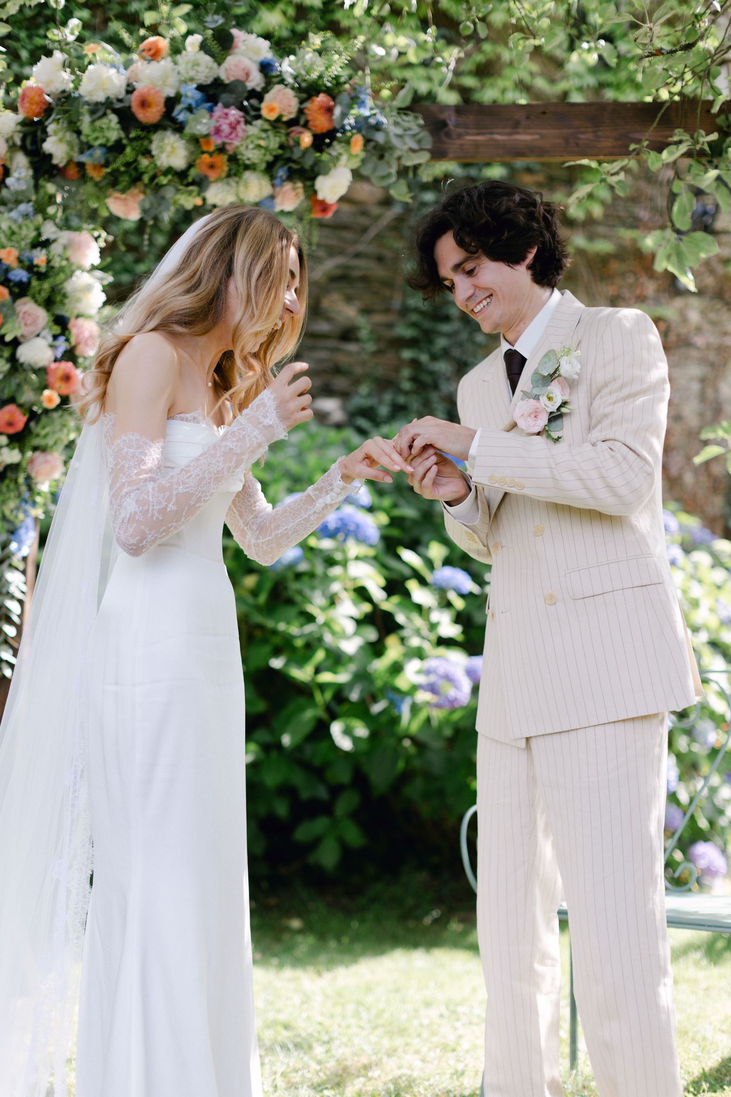 La mariée et le marié échangent des bagues lors de la cérémonie de mariage en plein air sous une arche florale colorée.