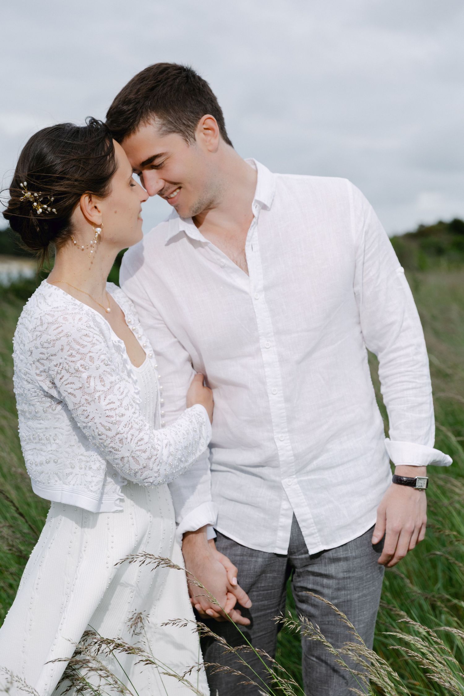 Un couple souriant en tenue blanche partage un moment romantique dans un champ vert luxuriant le jour de leur séance photo en couple.