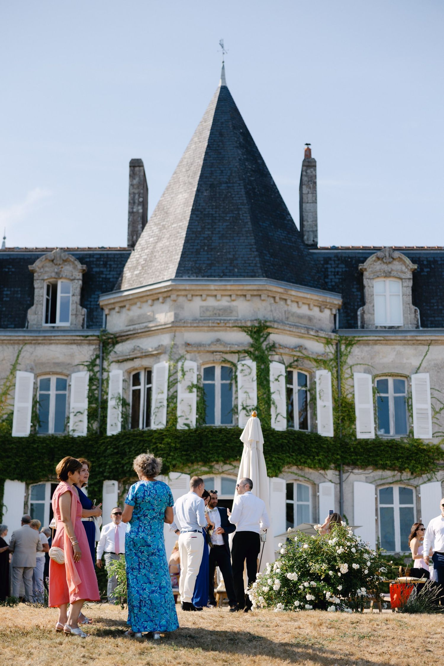 Guests mingle on the lawn of an elegant French château with a pointed slate roof on a sunny day.