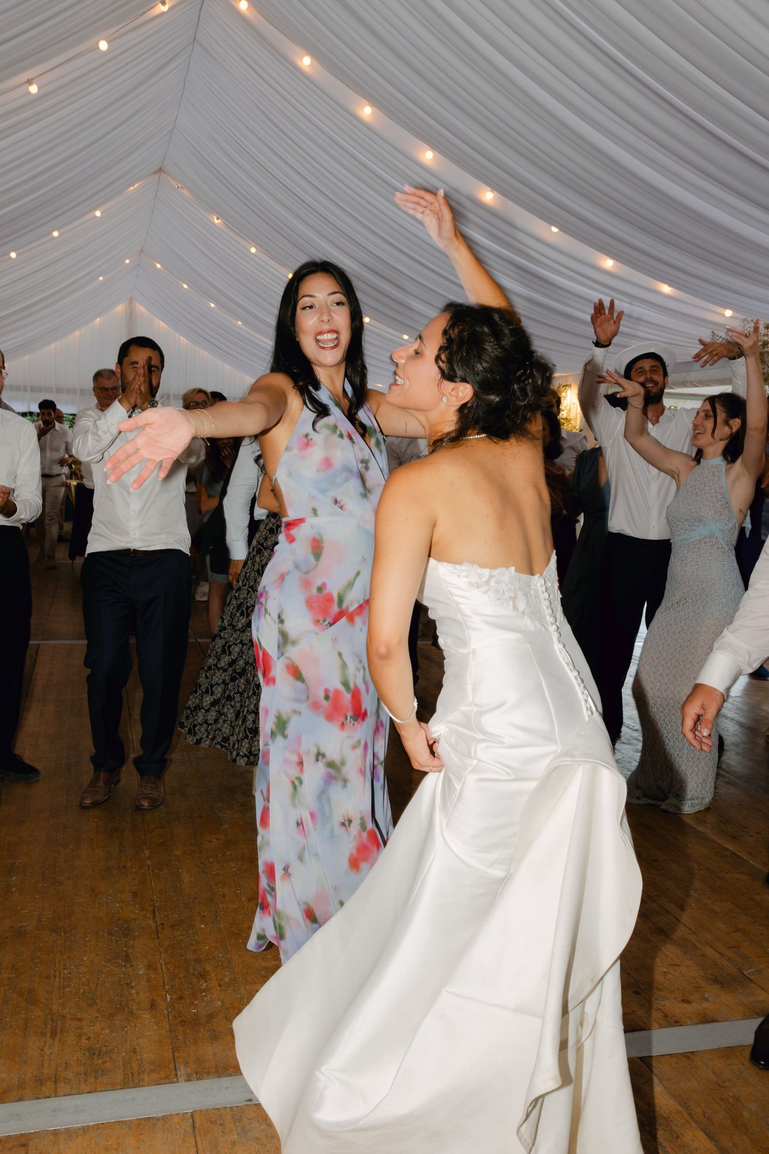 Bride in white gown dancing joyfully with guest in floral dress at elegant wedding reception under draped tent.