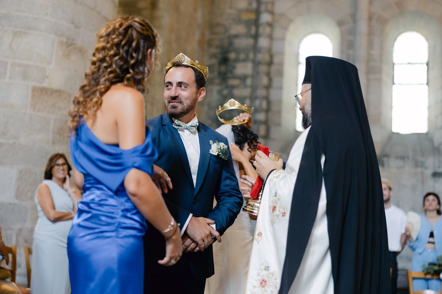 Orthodox wedding ceremony with groom in blue suit wearing crown, holding hands with bride, officiated by priest in black robes.
