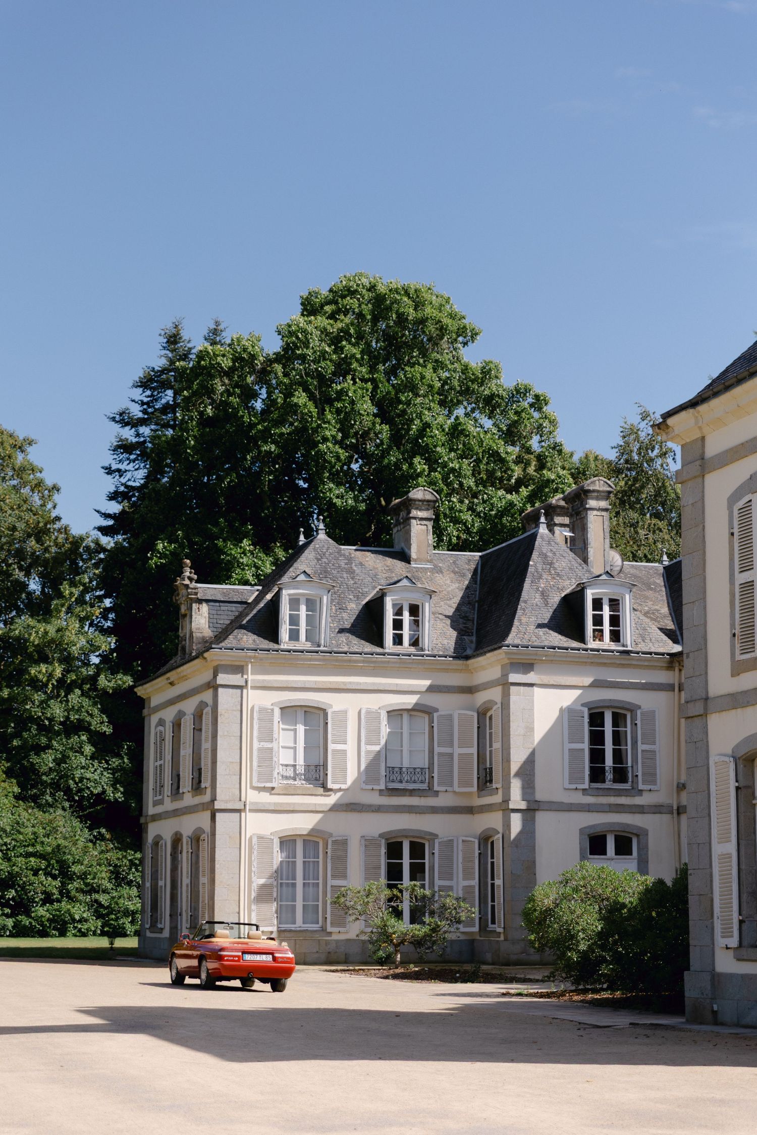 Une voiture rouge classique garée devant un élégant château français avec des fenêtres à volets et un toit en ardoise.