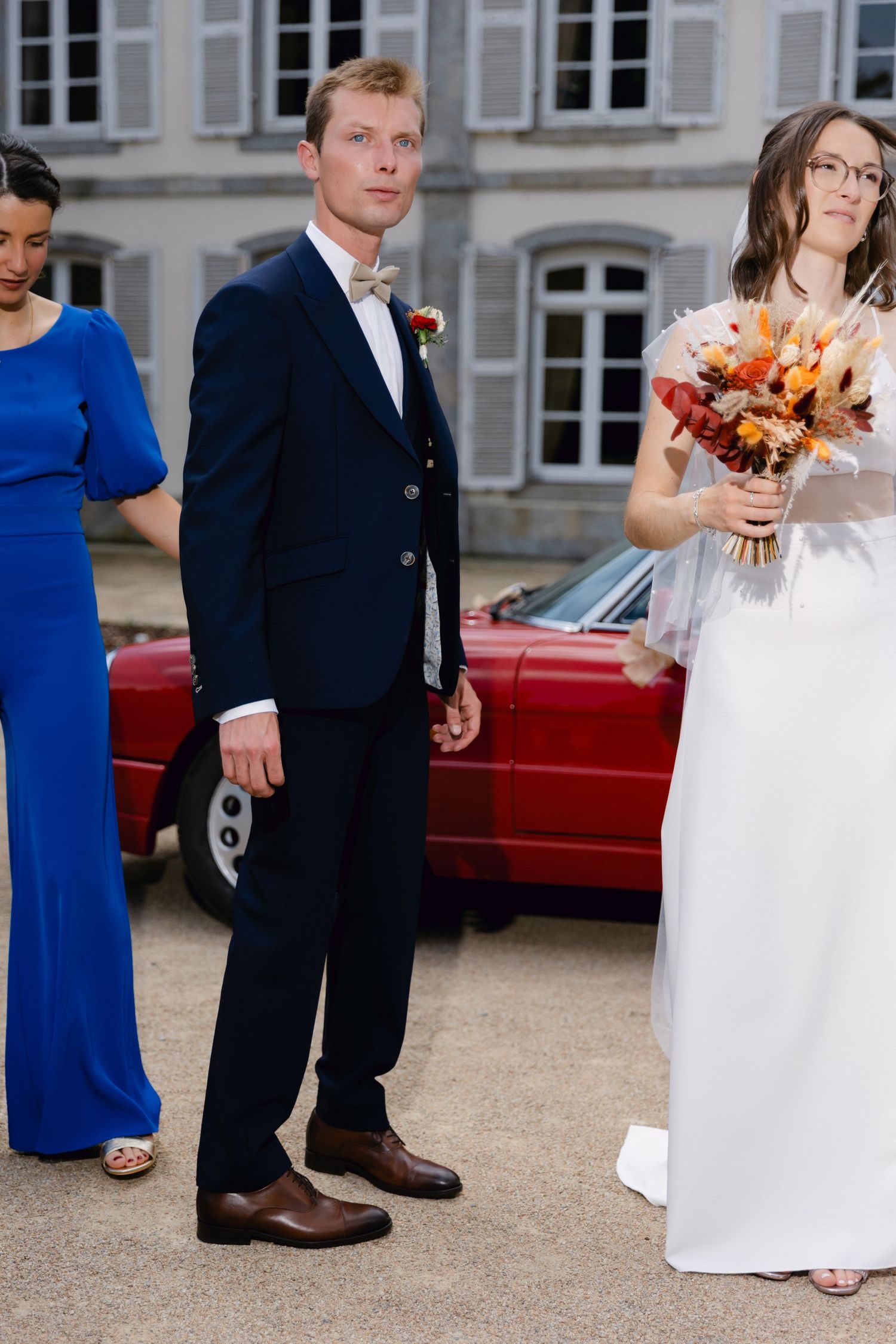 Le marié en costume bleu marine se tient à côté de la mariée en robe blanche tenant un bouquet d'automne près de la voiture rouge lors d'un