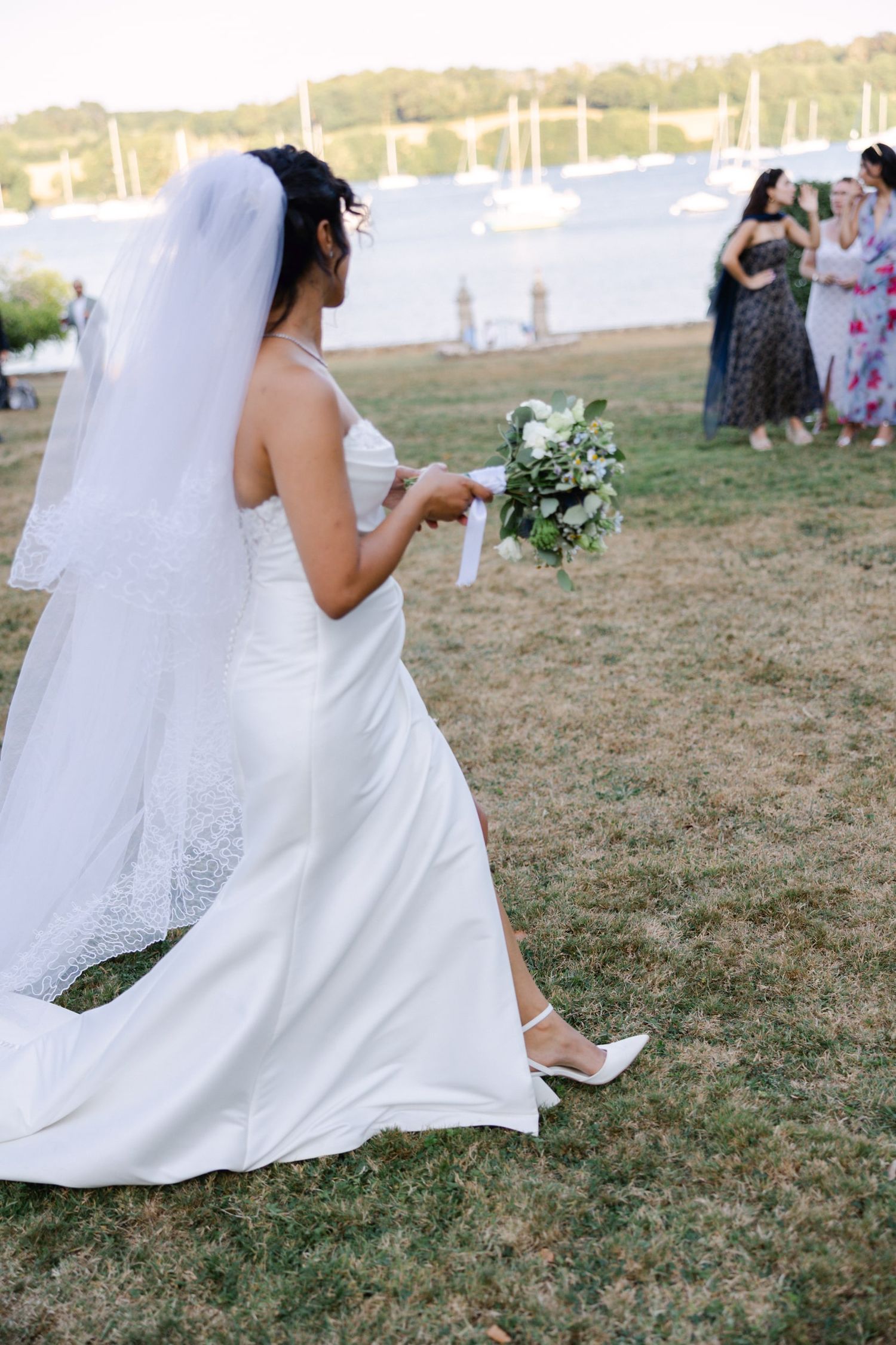 Bride in white gown and veil holds green bouquet outdoors near waterfront, facing guests on grass.