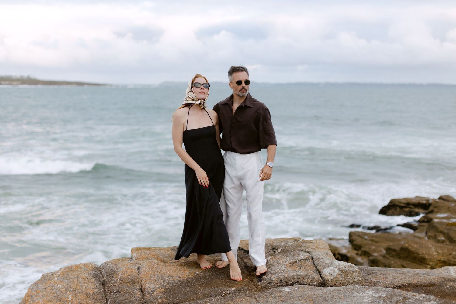 Couple posant sur un rivage rocheux avec des vagues d'océan, femme en robe noire et homme en pantalon blanc et chemise marron.