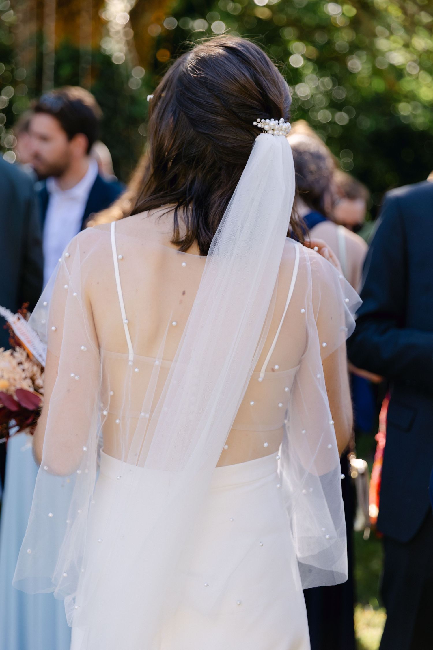 Mariée en robe de mariée en tulle orné de perles avec voile, vue de derrière lors d'une cérémonie en plein air.