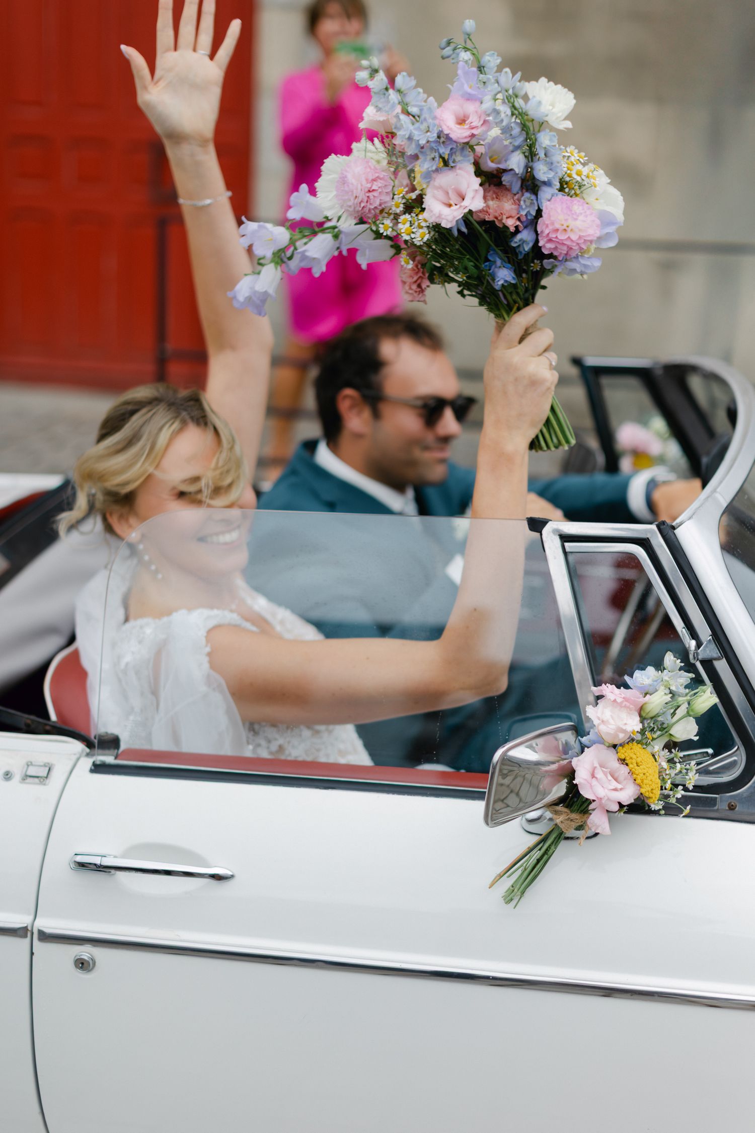 Une mariée jubilante lève un bouquet pastel tout en conduisant une voiture décapotable blanche avec le marié après le mariage.
