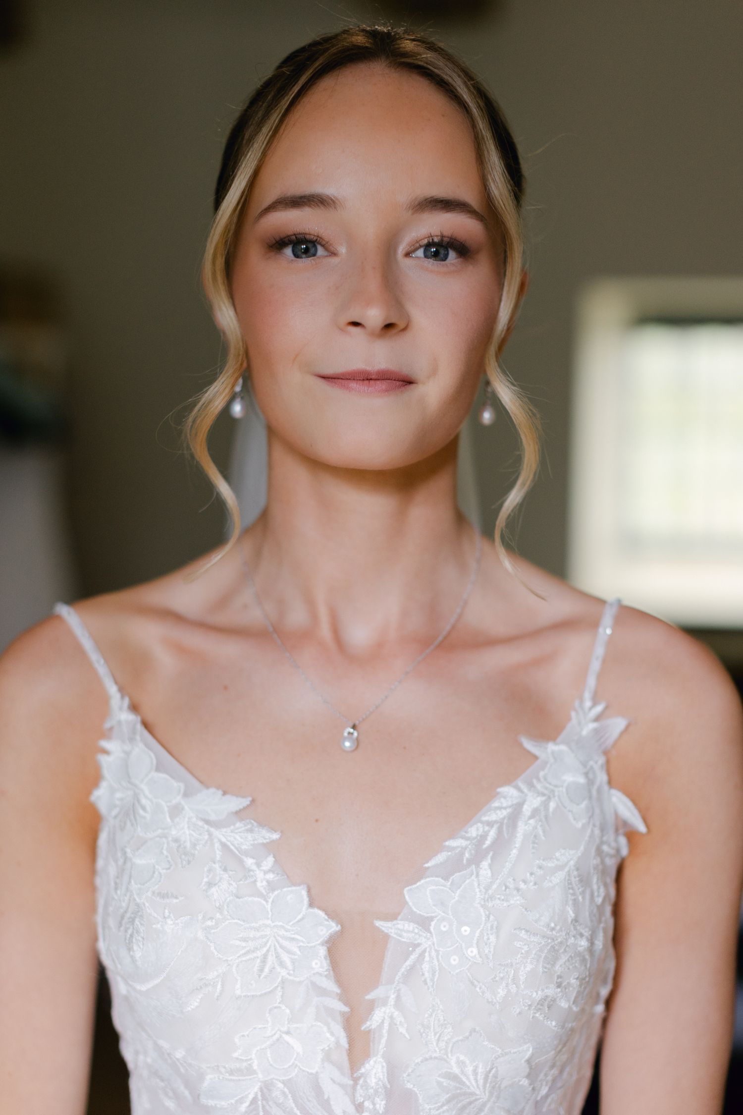 Mariée en robe de mariée à bretelles spaghetti en dentelle avec boucles d'oreilles en perles et collier souriant doucement.