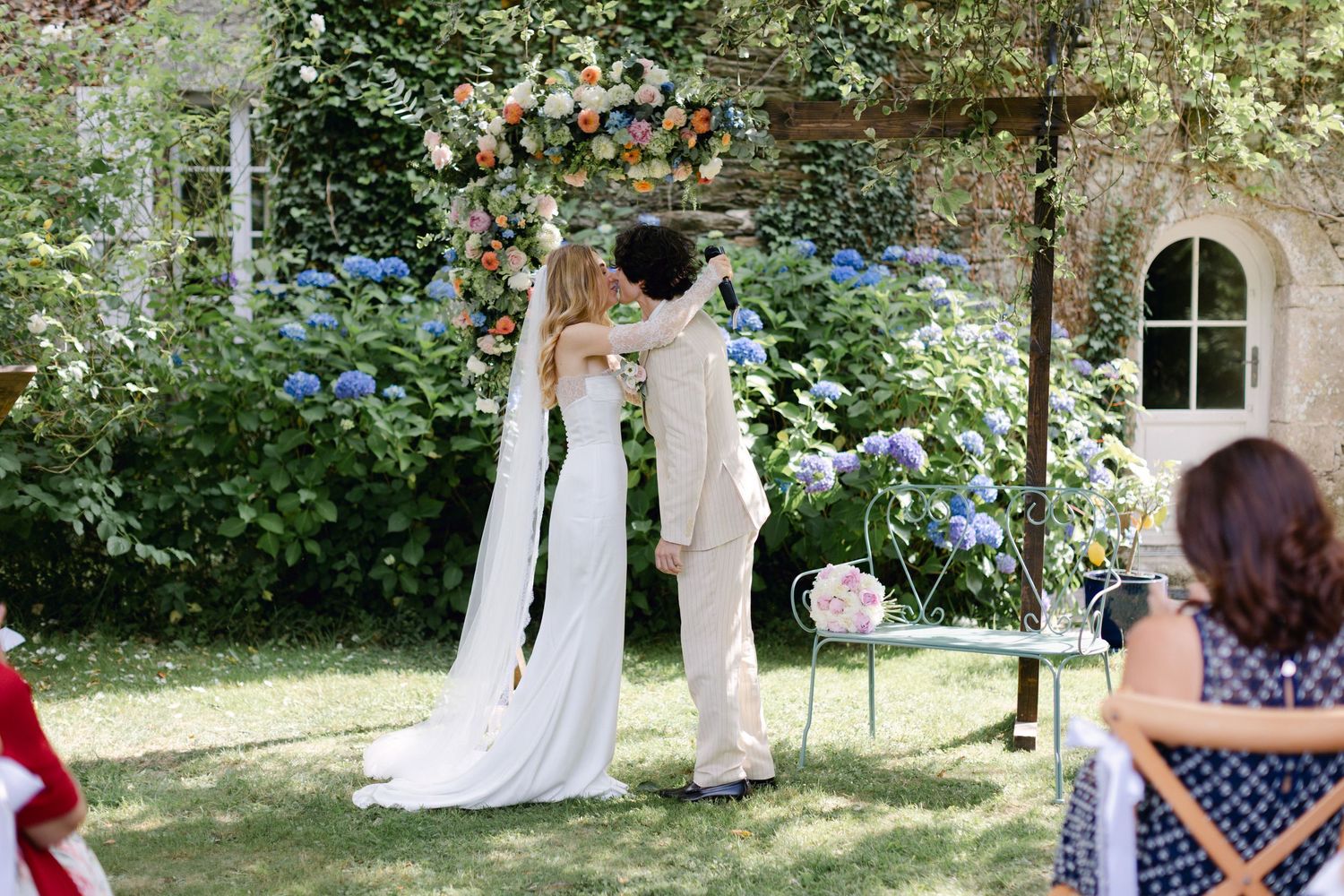 La mariée et le marié échangent leurs vœux sous une arche florale vibrante lors d'une cérémonie de mariage dans un jardin en plein air.
