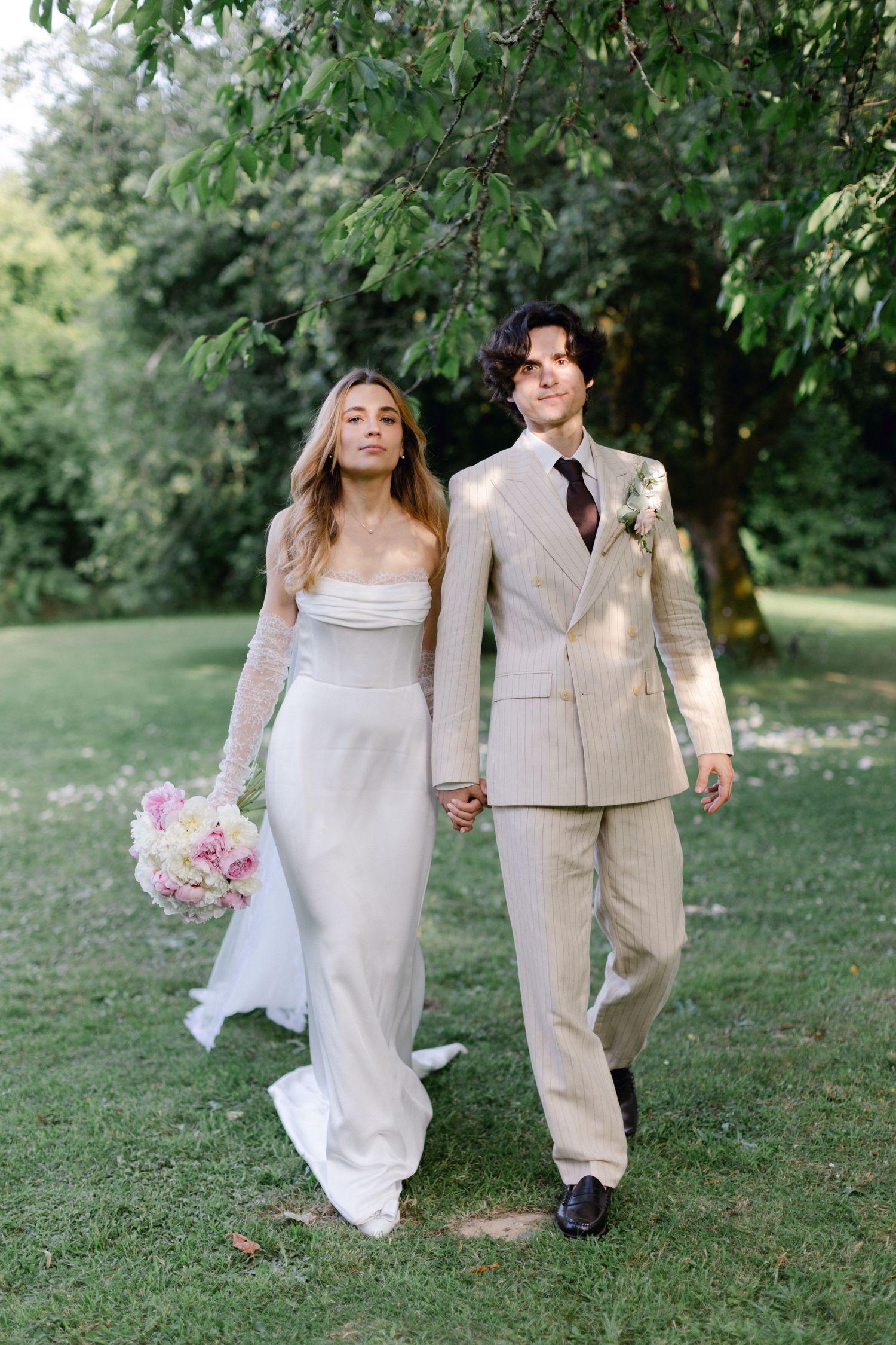 Les jeunes mariés souriants marchent main dans la main dans un jardin ensoleillé, la mariée tenant un bouquet rose, le marié en costume.