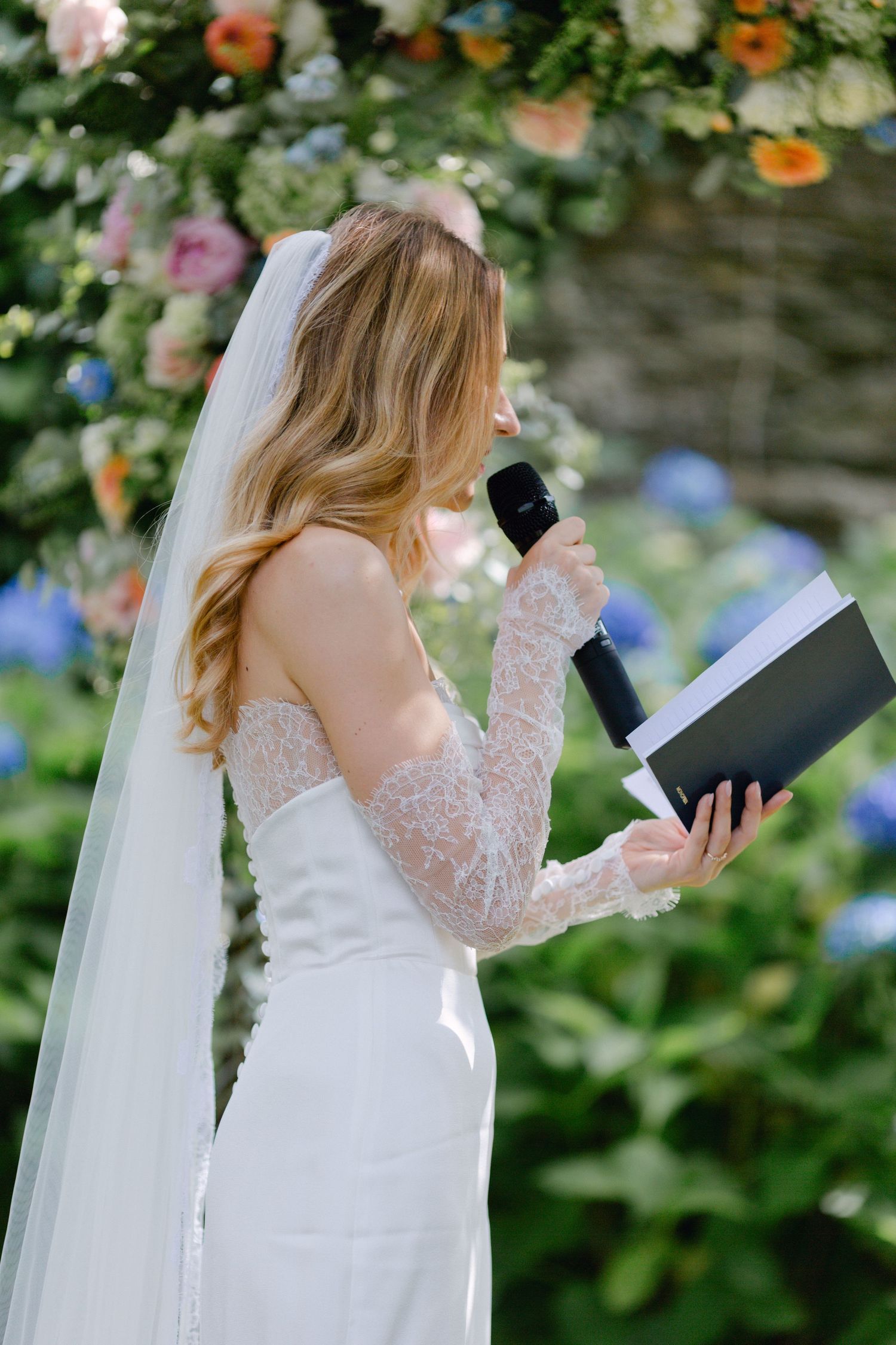Une mariée élégante en dentelle à épaules dénudées lit les vœux de mariage dans le microphone sous une arche florale colorée.