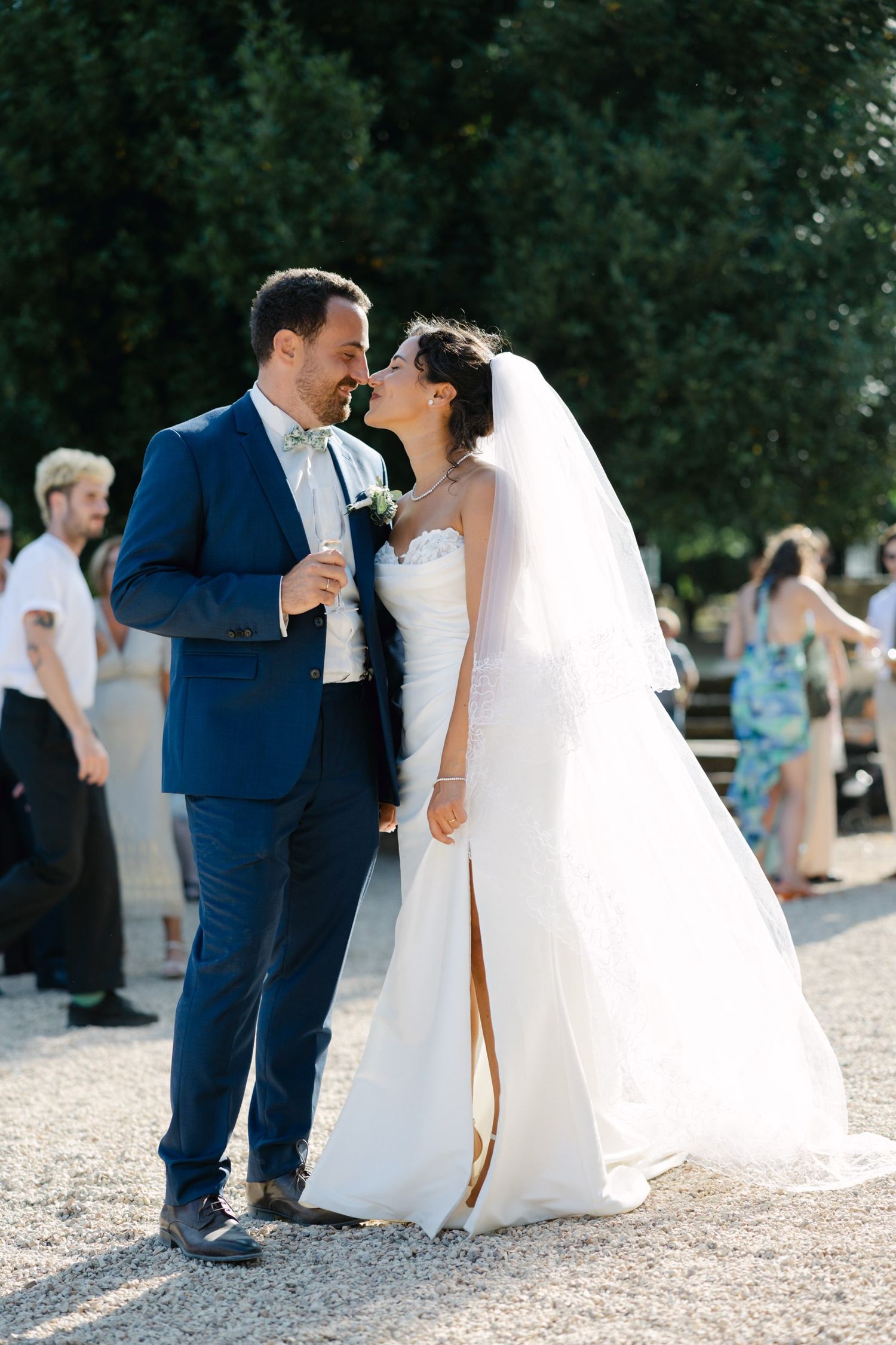 Bride in white gown and veil kissing groom in blue suit outdoors at sunny wedding reception.