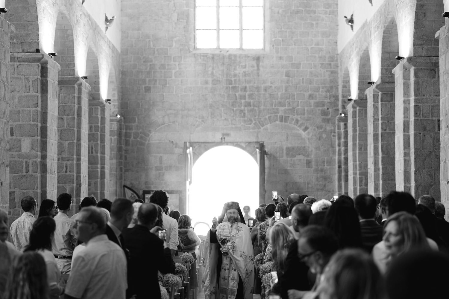 Black and white photo of a wedding ceremony inside a historic stone church with arched columns and bright altar light.