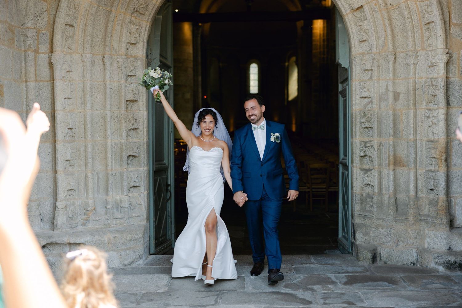 Joyful newlyweds exit stone church holding hands, bride raises bouquet in celebration wearing white gown, groom in navy suit.