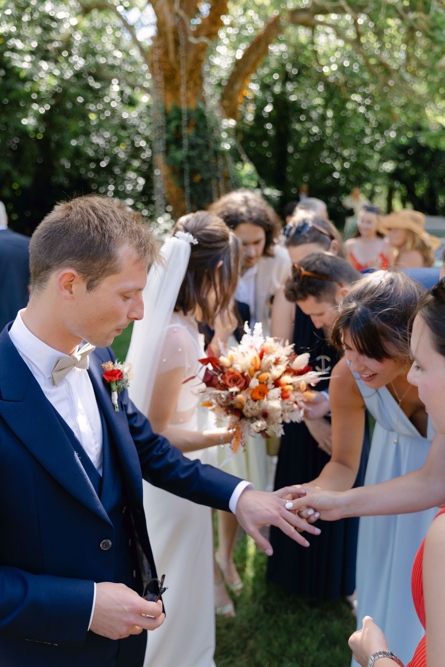 Le marié en costume bleu marine montre sa bague à ses invités lors d'un cocktail de mariage en plein air.