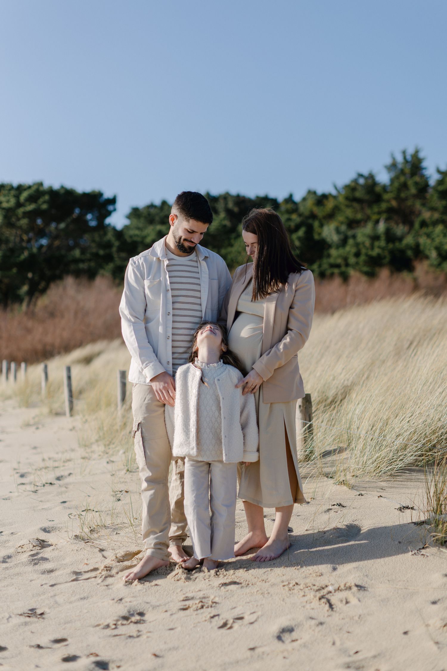 Photo de maternité familiale avec père, fille et mère enceinte debout ensemble sur des dunes de sable ensoleillées.