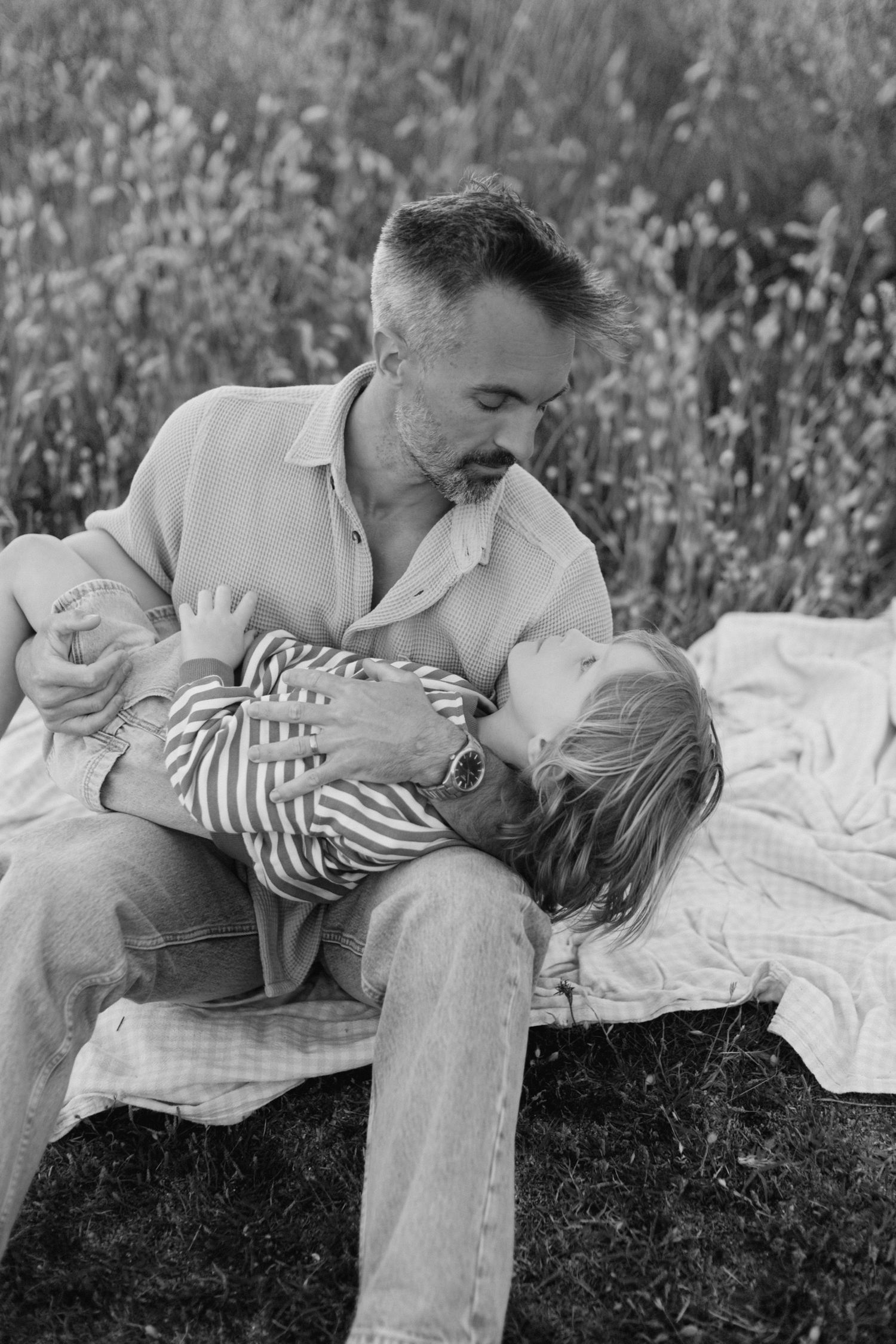 Photo en noir et blanc d'un père assis à l'extérieur berçant son jeune enfant sur une couverture dans un champ.