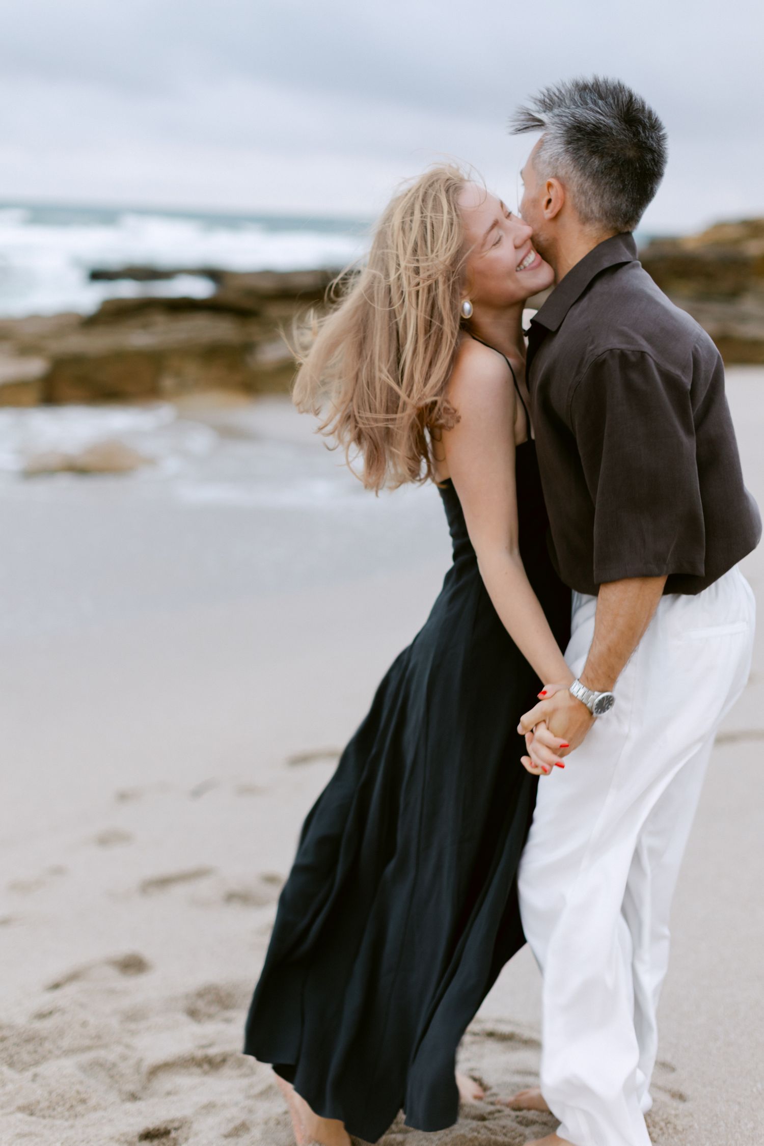Un couple partage un baiser romantique sur une plage de sable, la femme en robe noire avec des cheveux qui soufflent dans le vent.
