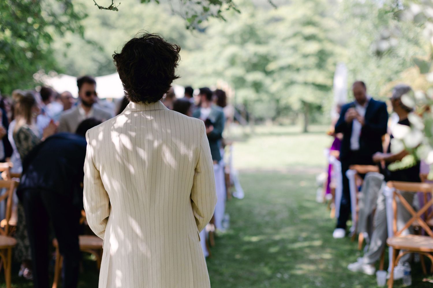 Personne en costume rayé crème regardant une cérémonie de mariage en plein air par derrière parmi les invités assis.