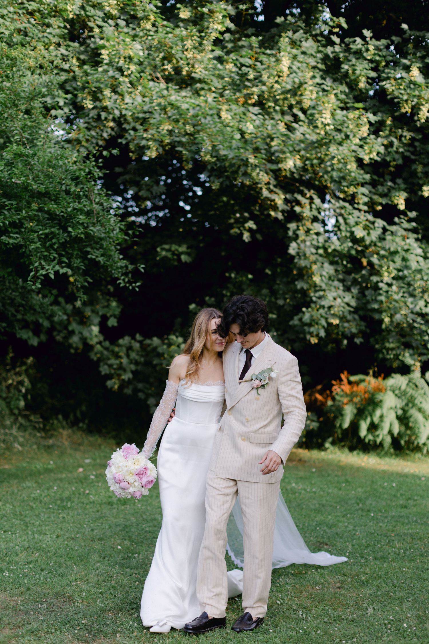 Couple de mariage se promenant sur une pelouse verte, mariée en robe blanche sans bretelles tenant un bouquet rose, marié en costume.