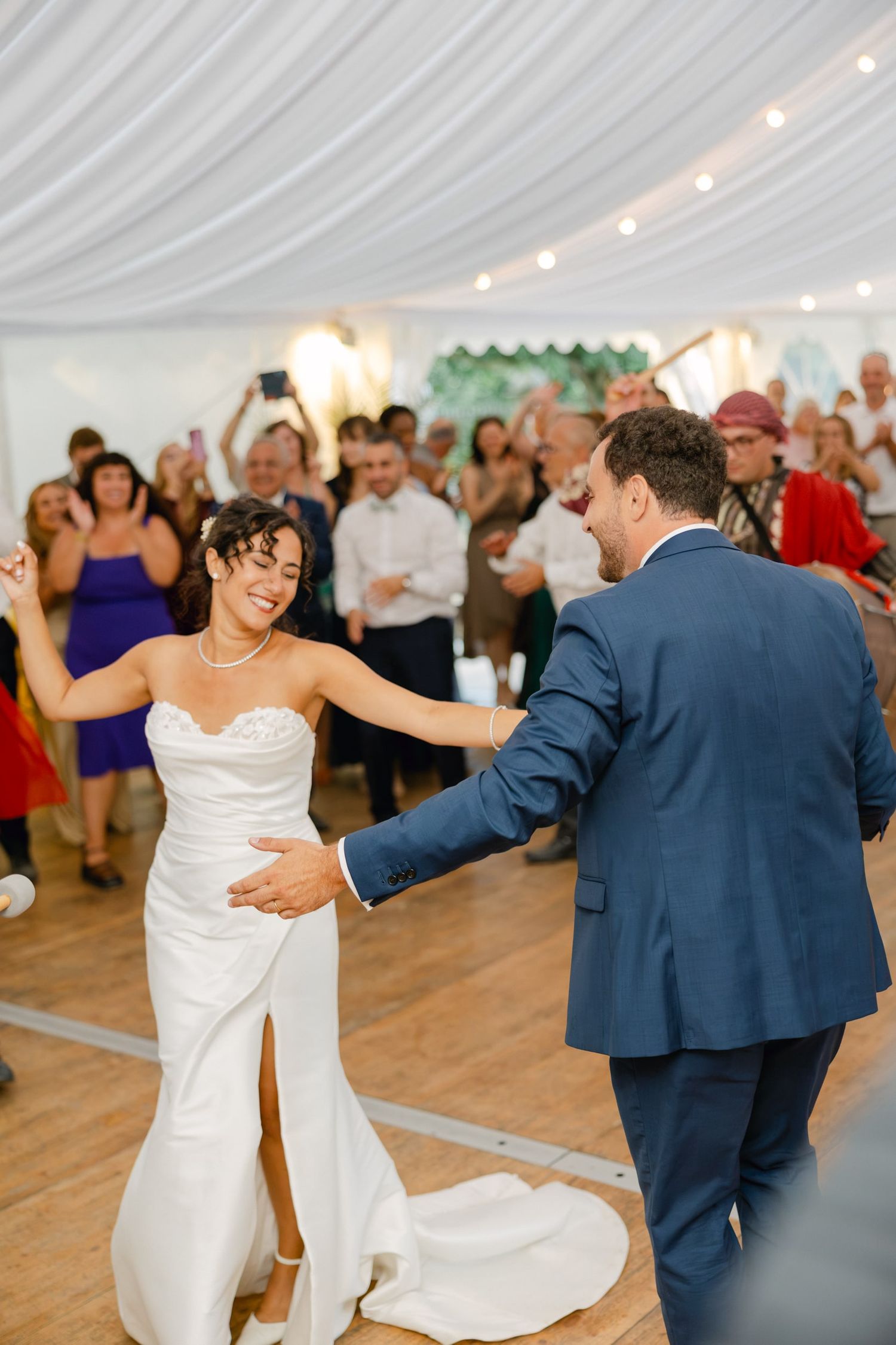 Bride and groom share their first dance at wedding reception, guests cheering in background under white draped tent.