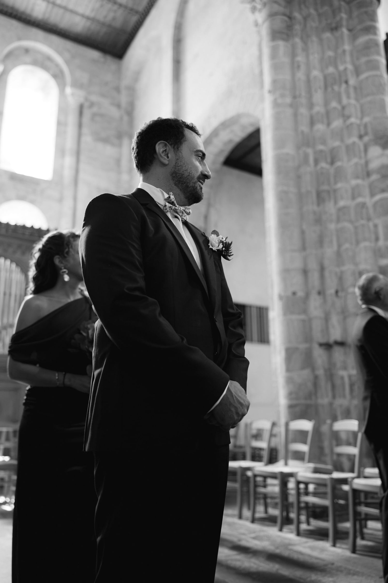 Groom in black tuxedo and bow tie smiling at altar in stone church during wedding ceremony, black and white photo.