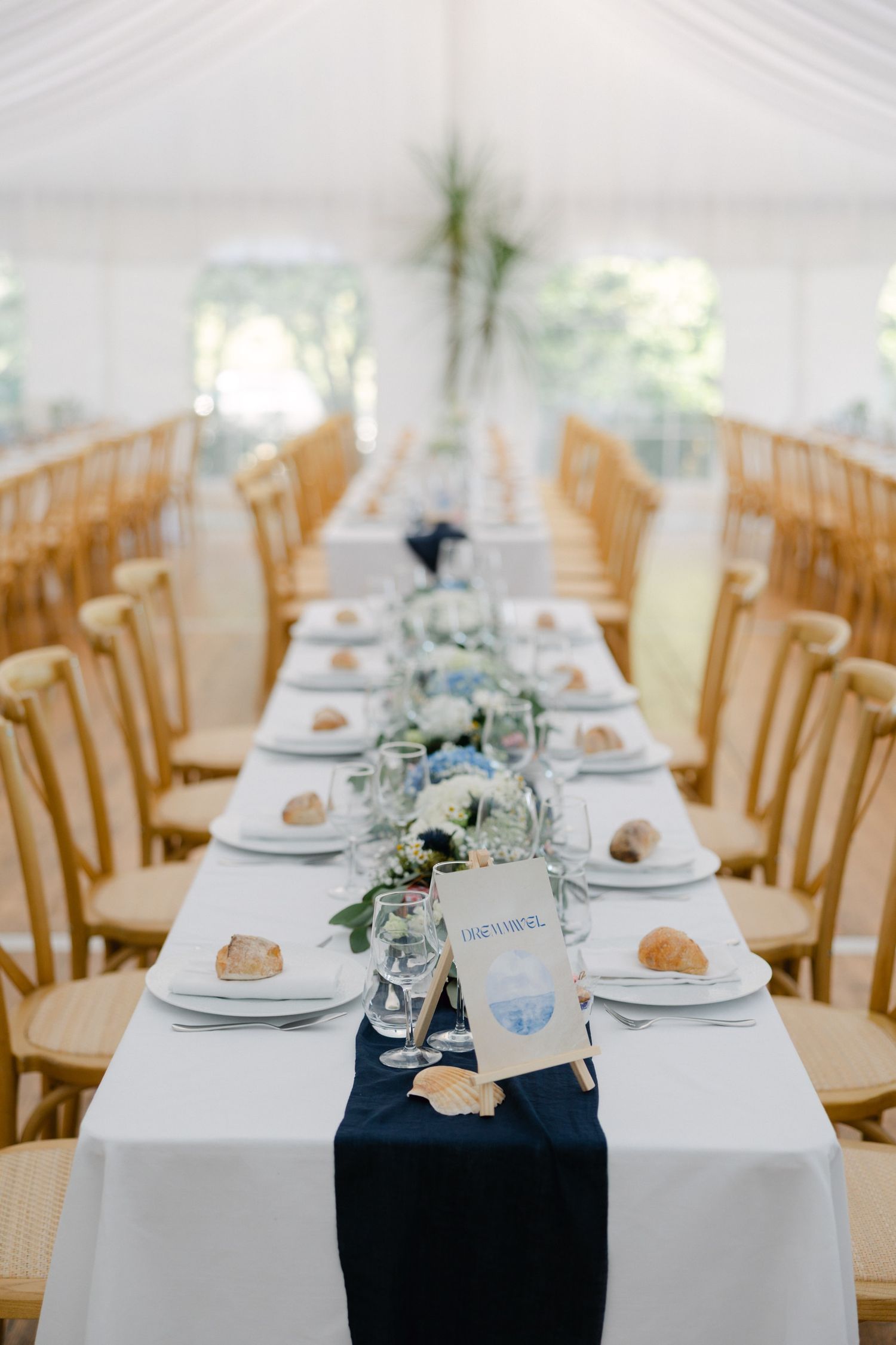 Elegant wedding reception table with navy blue runner, white linens, wooden chairs, and floral centerpieces in a white tent.