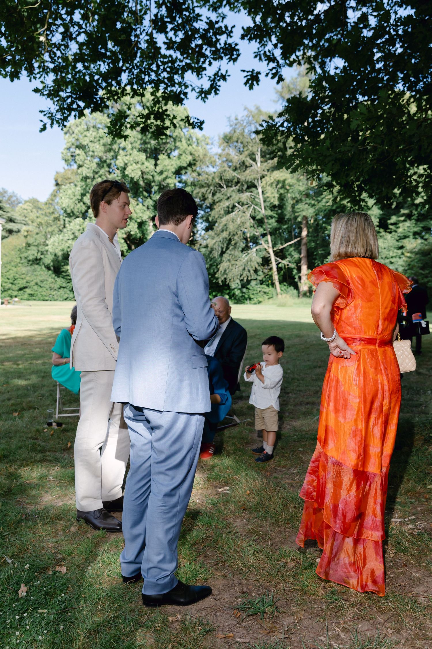 Les invités en tenue formelle se rassemblent à l'extérieur sous les arbres lors d'une réception de mariage d'été par une journée ensoleillée