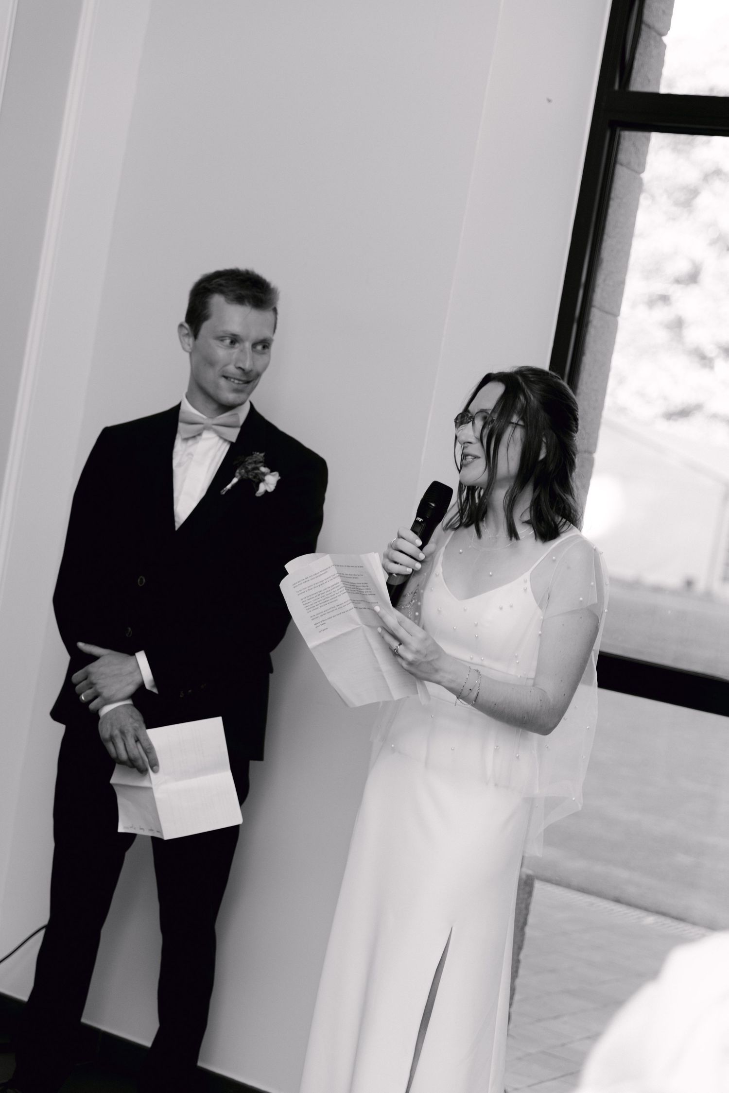Photo en noir et blanc d'une mariée lisant des vœux à haute voix lors d'une cérémonie de mariage tandis que le marié sourit à côté d'elle.