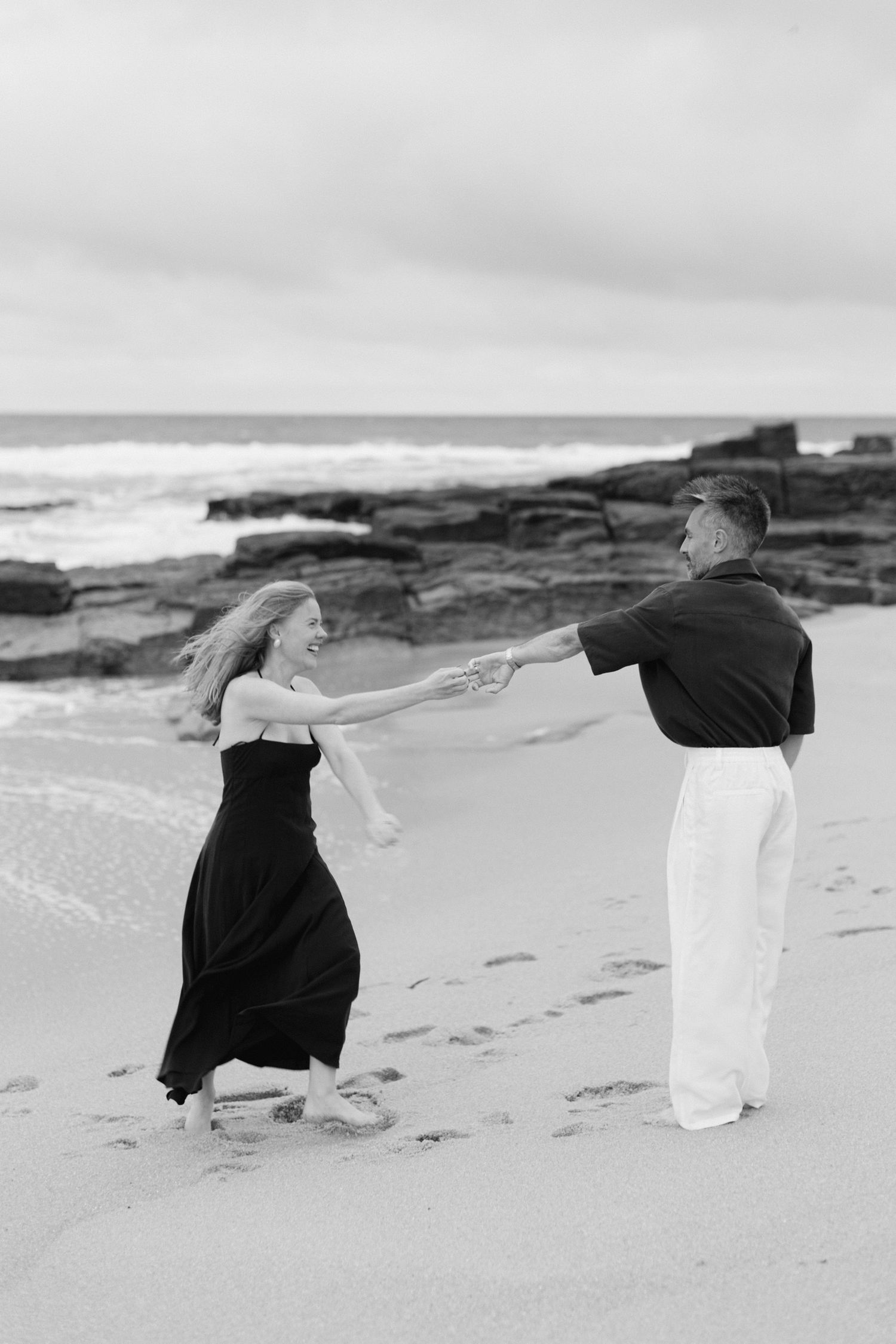 Photo en noir et blanc d'un couple dansant joyeusement sur une plage de sable avec des rives rocheuses et des vagues de l'océan.