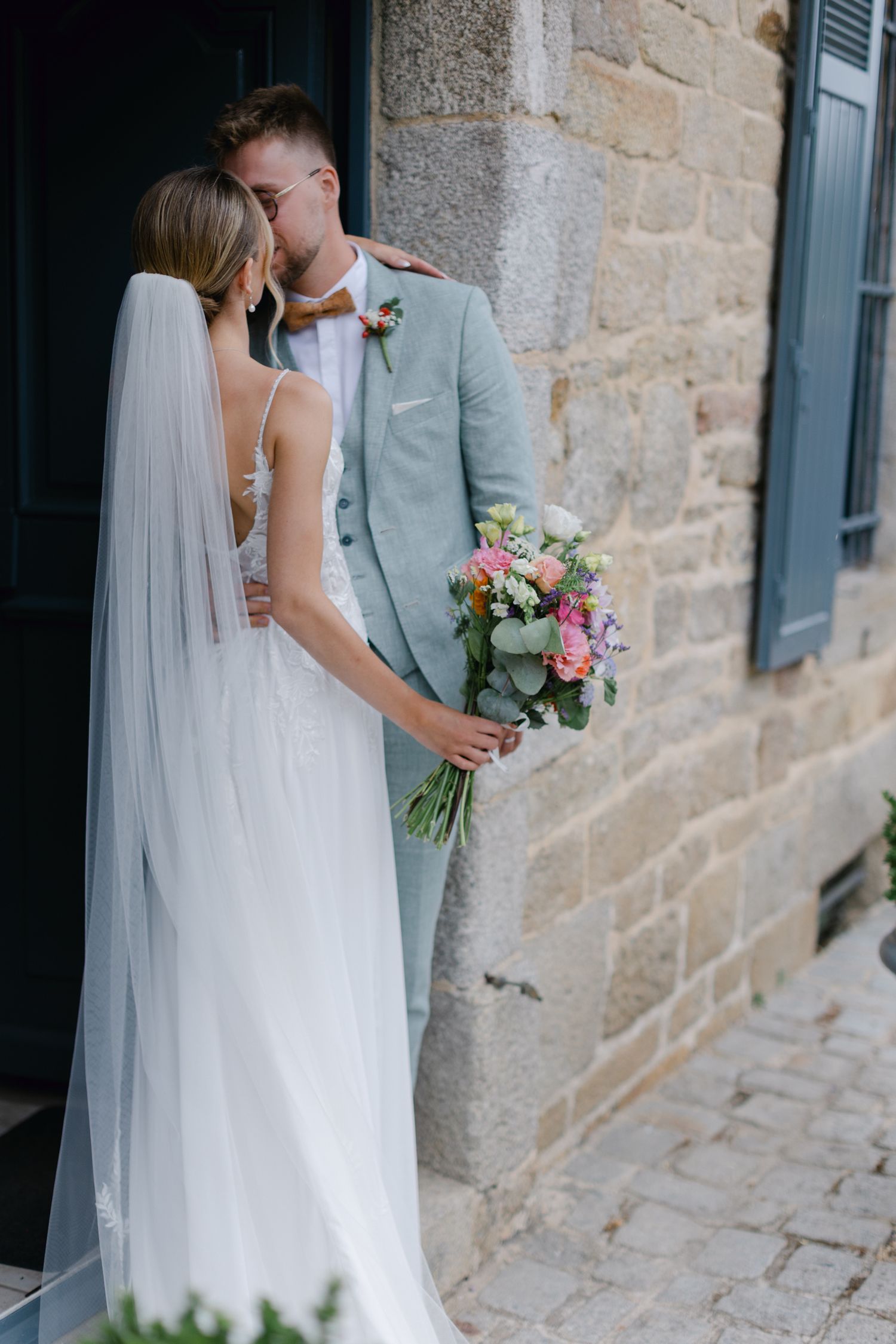 La mariée et le marié partagent un baiser intime à côté d'un bâtiment en pierre rustique tenant un bouquet de mariage coloré.